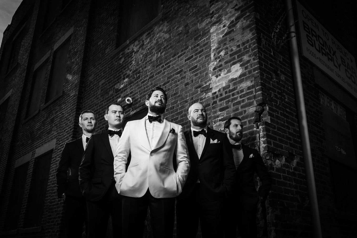 Black and white photo of a wedding party in formal attire standing against a brick wall.