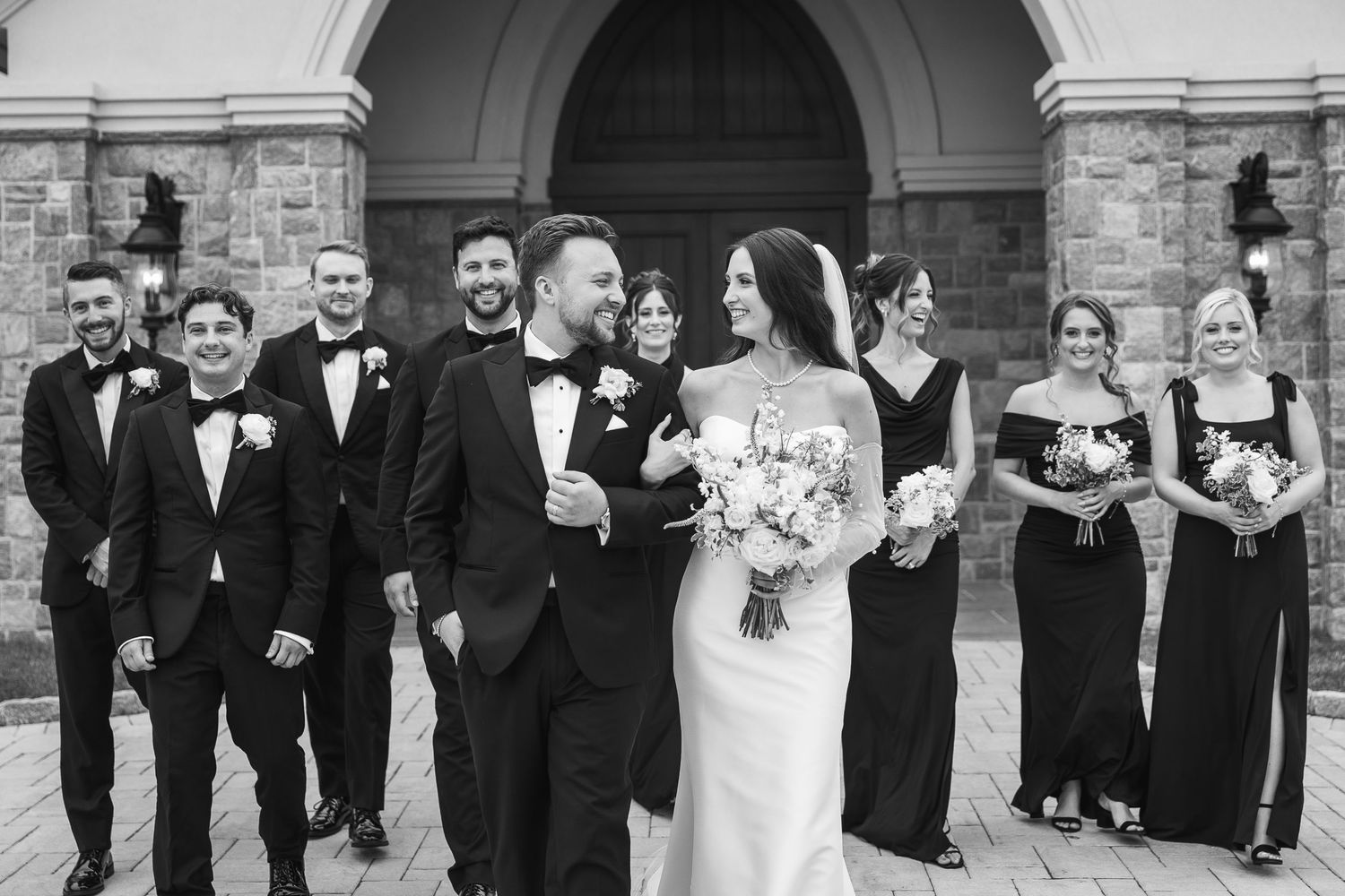 Wedding party in formal attire poses under stone archway for group photos.