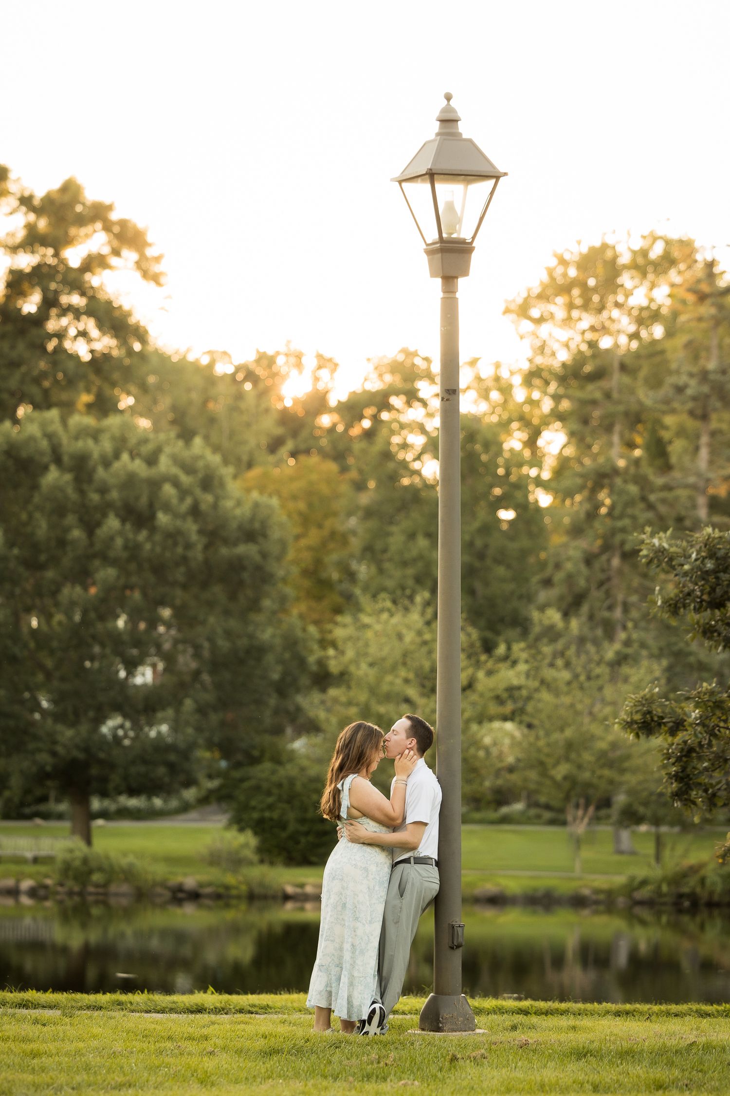 Couple kissing under a vintage lamp post in a lush green park at golden hour sunset.