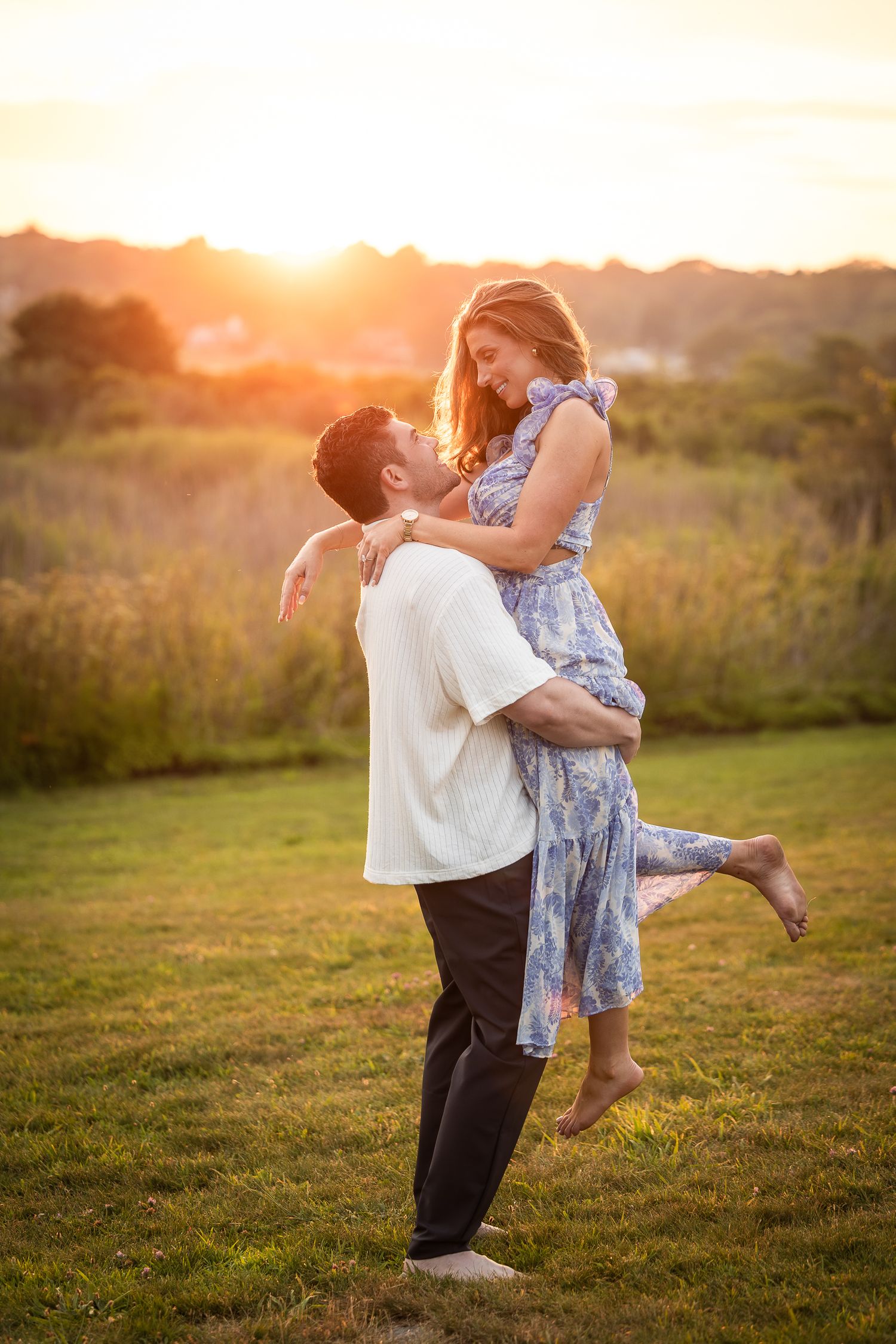 A romantic couple embraces in a grassy field during golden hour sunset as she is lifted off her feet.