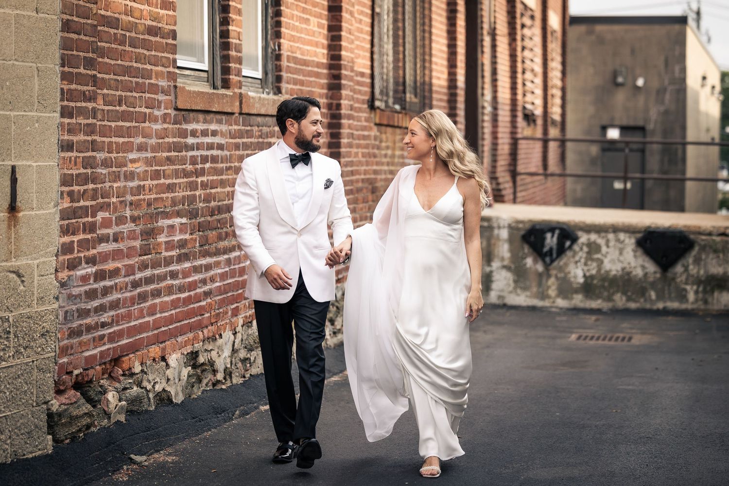 Elegant couple walking along brick wall exterior during wedding photoshoot.