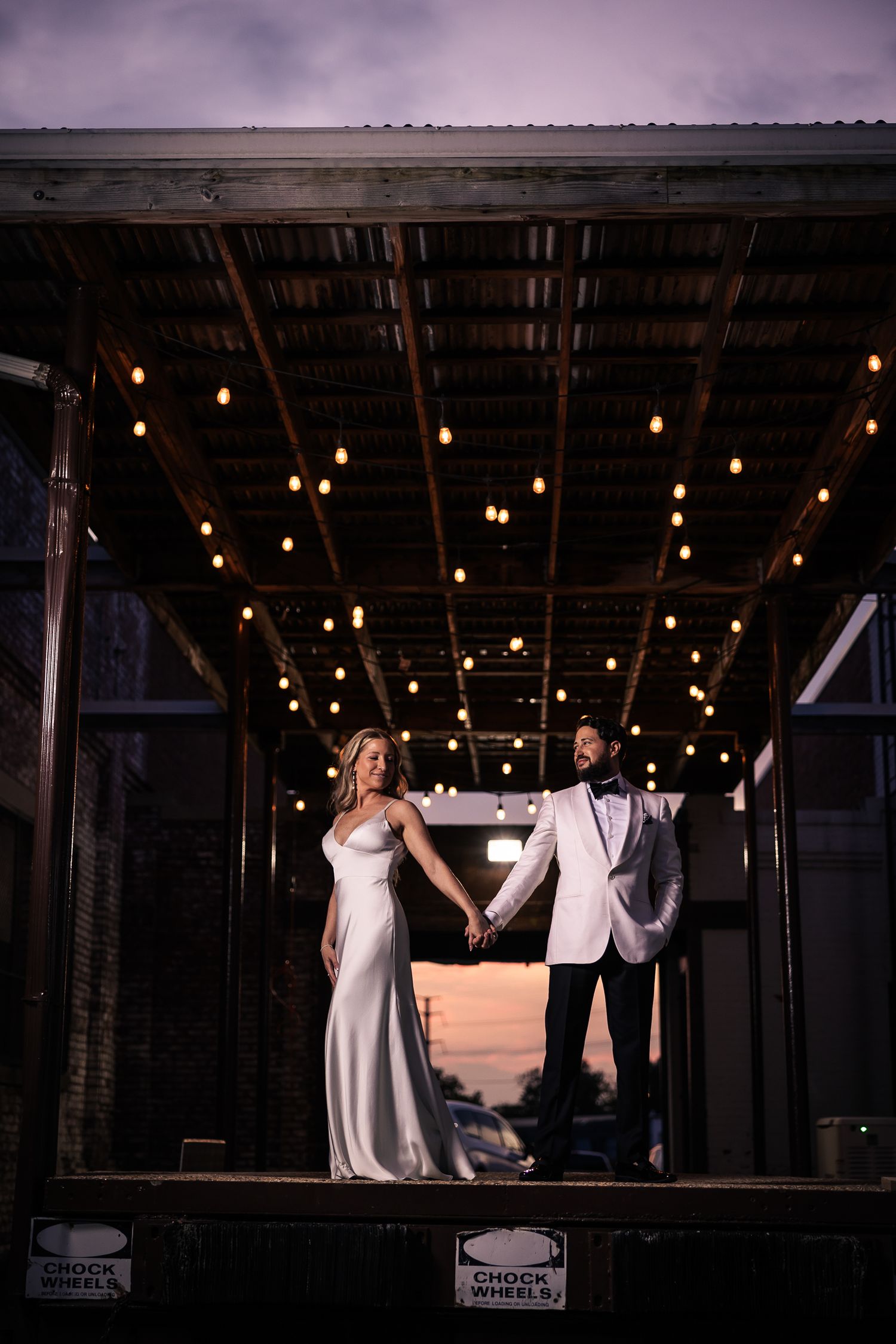A romantic silhouette of a couple dancing under string lights in an industrial venue during their wedding celebration.