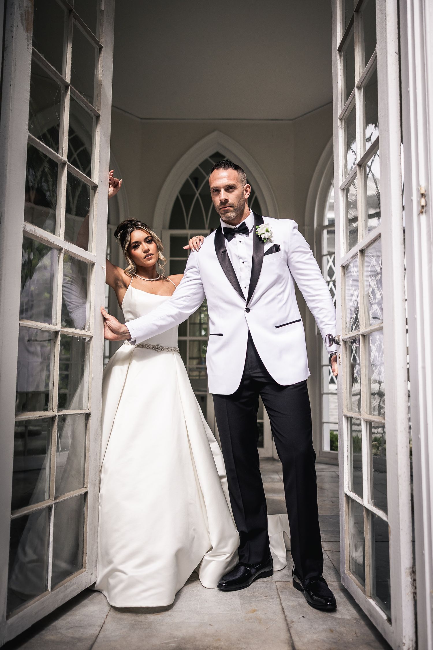 Wedding party poses playfully in elegant white and black formal attire along a glass-windowed hallway.