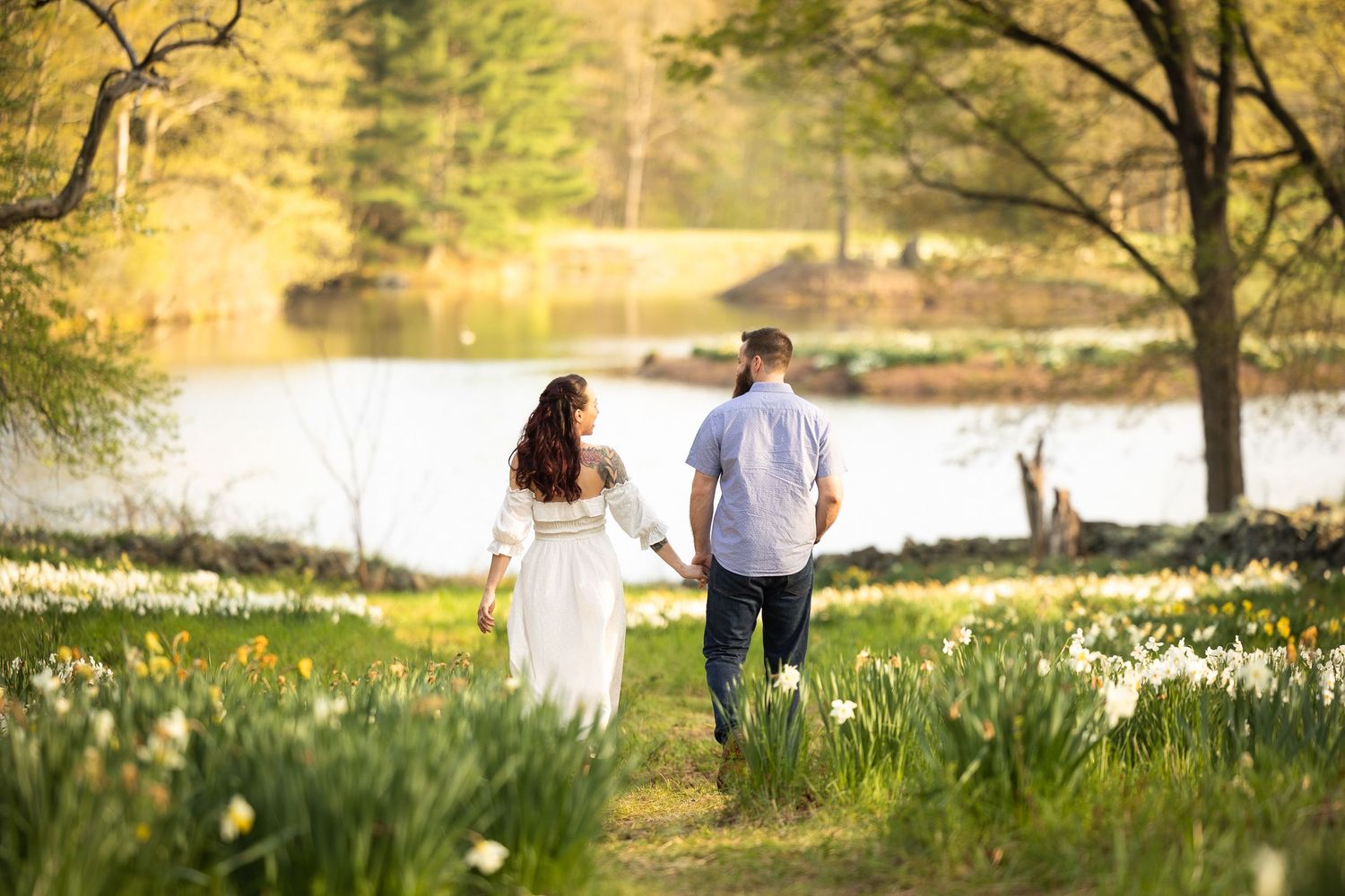 Two people walking together on a spring path by a lake surrounded by blooming daffodils in golden afternoon light.