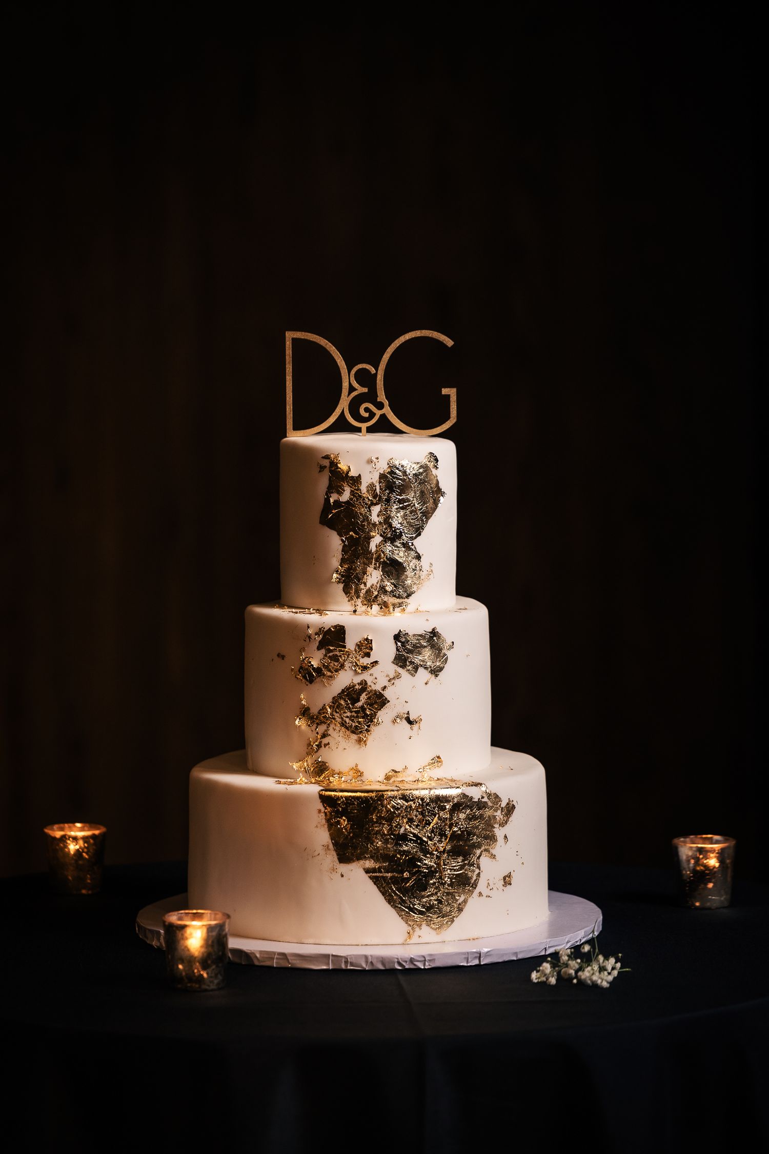 A white wedding cake decorated with metallic black butterflies photographed in a dark setting with glowing candlelight.