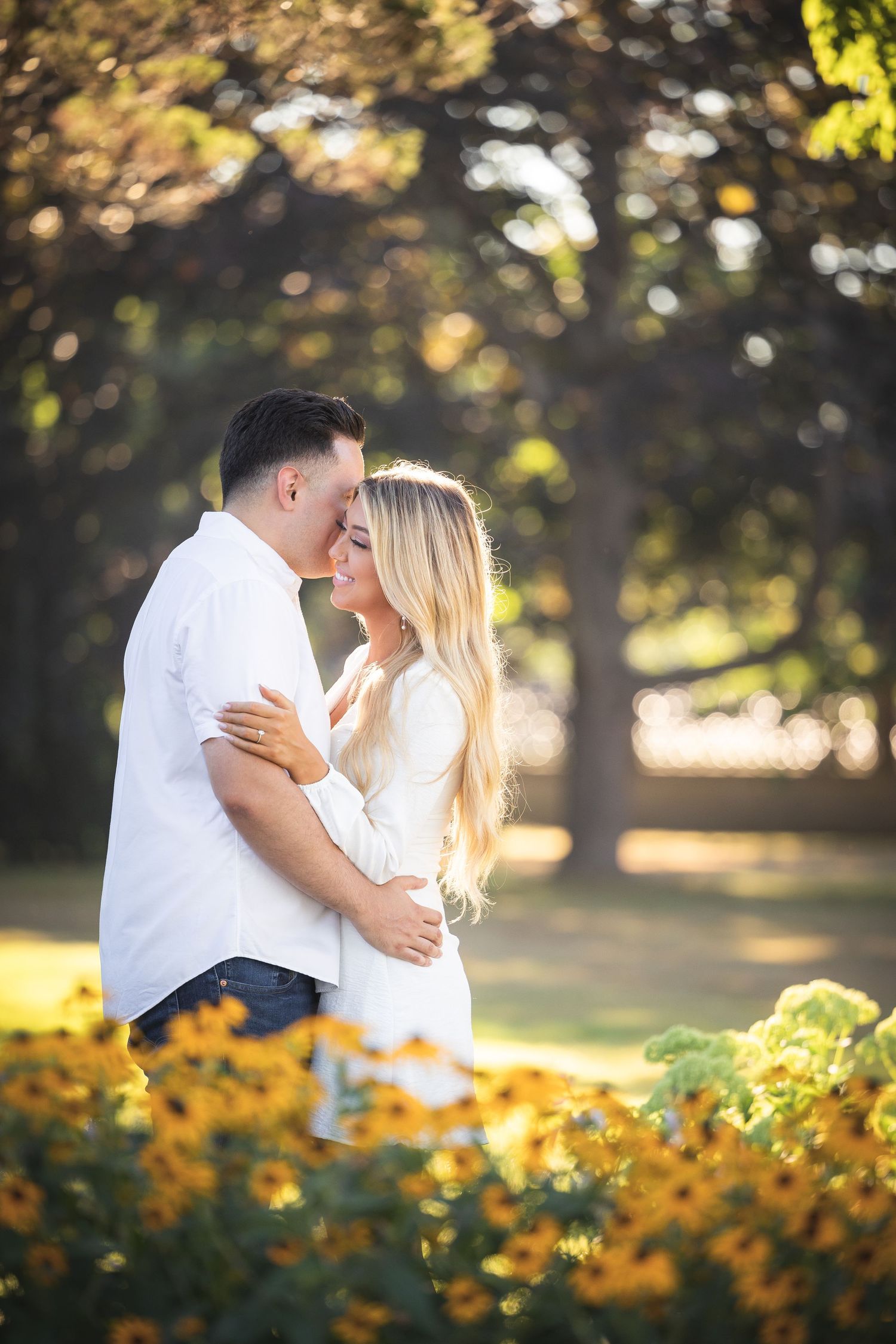 A romantic couple embraces in a sunlit park with golden flowers in the foreground during a summer evening.