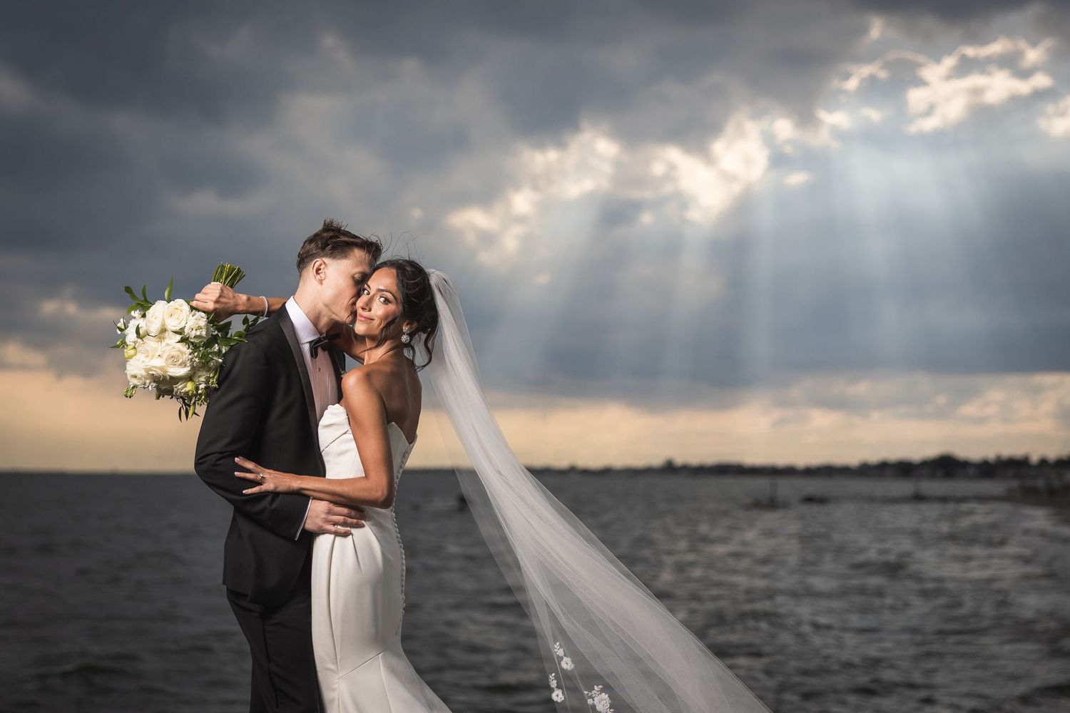 Dramatic sunset wedding photos captured against stormy sky over water.