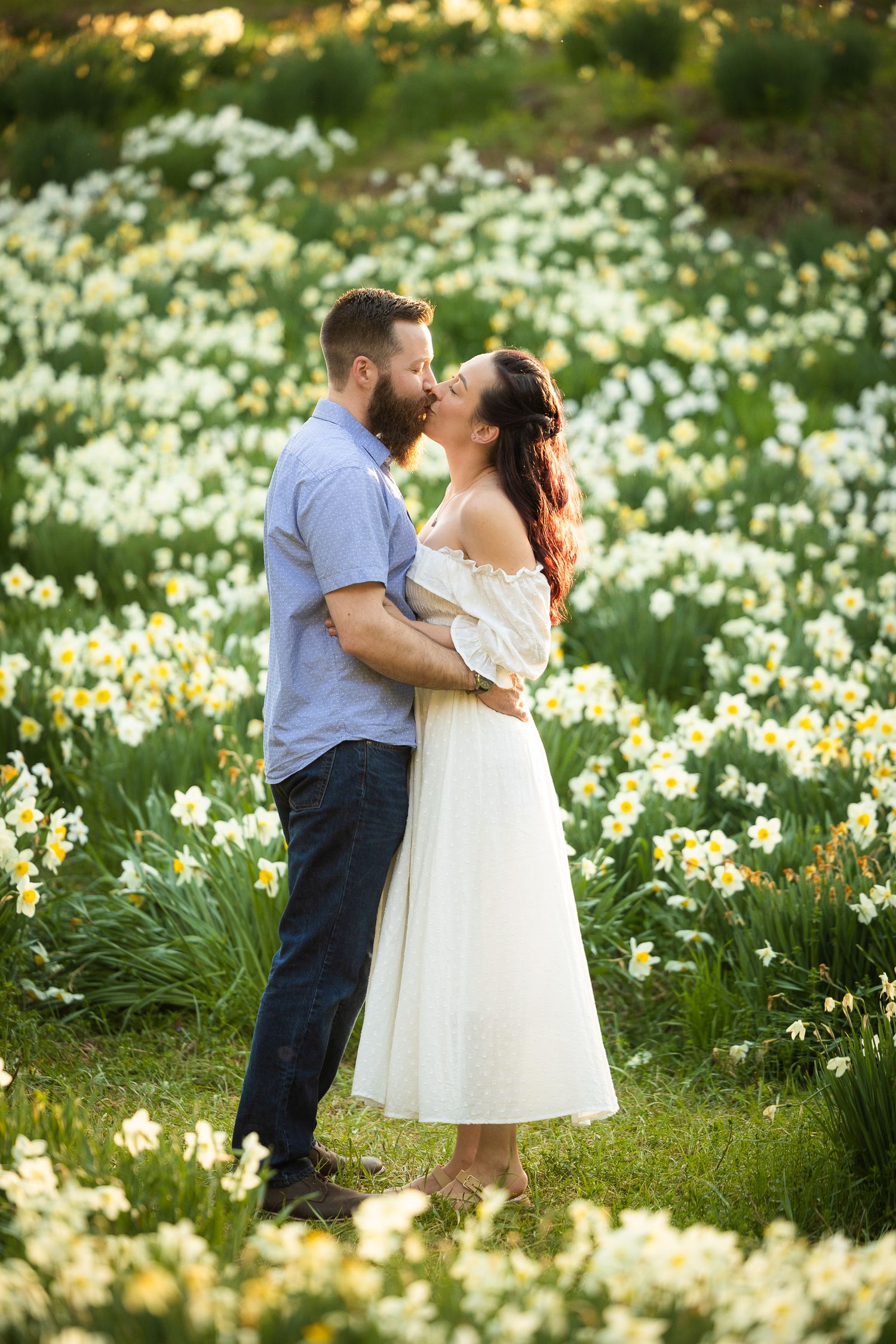 A romantic couple shares tender moments in a blooming spring meadow filled with white daffodils at sunset.