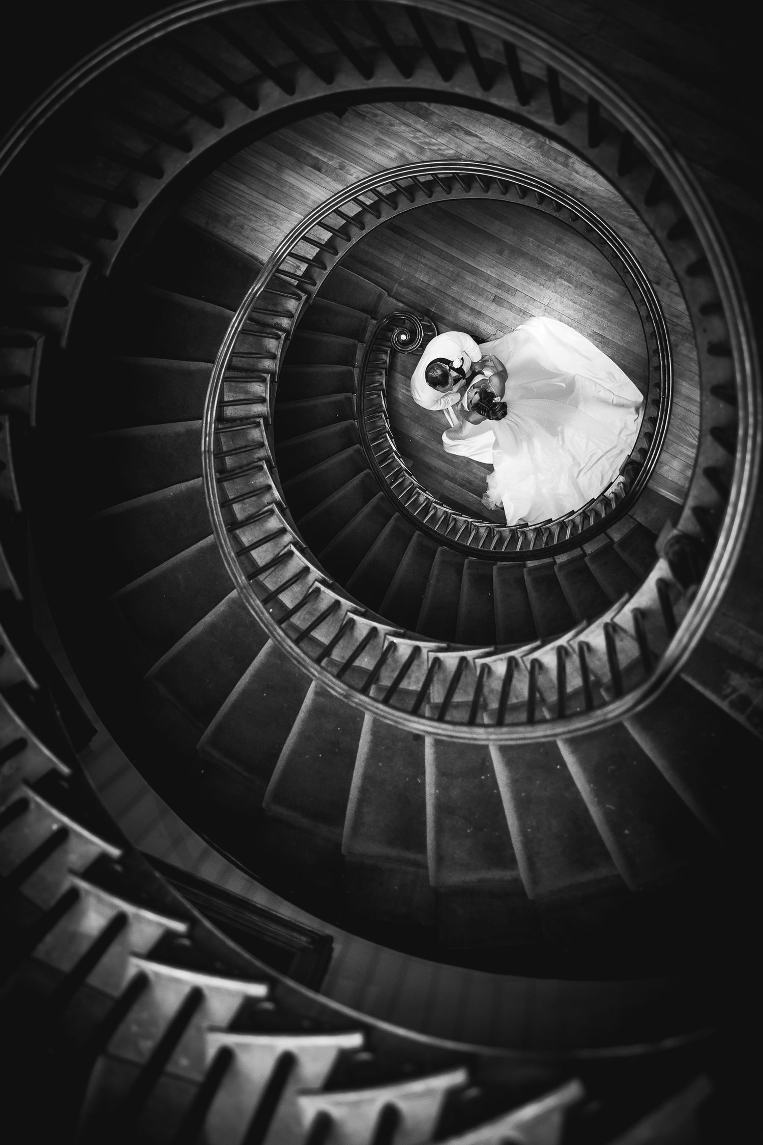 Black and white spiral staircase wedding photo with dramatic lighting from above.