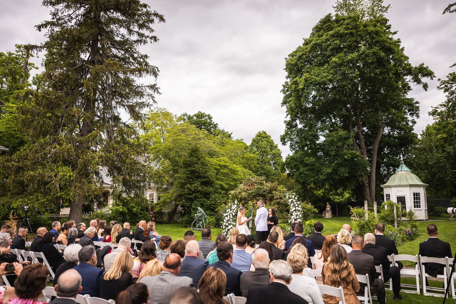 Panoramic view of outdoor wedding ceremony with guests seated under tall trees.