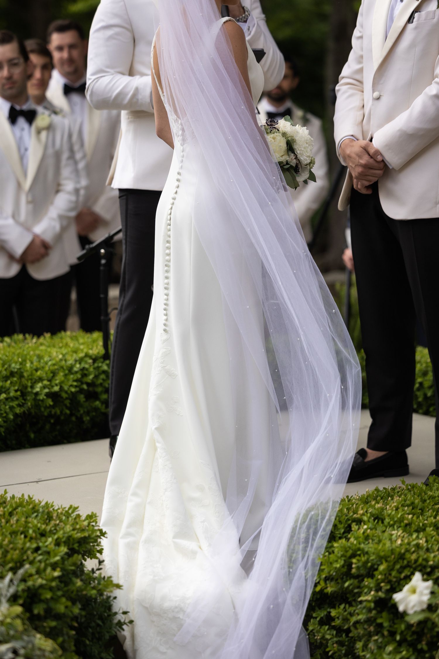 Close-up details of flowing white wedding dress and veil against garden backdrop.