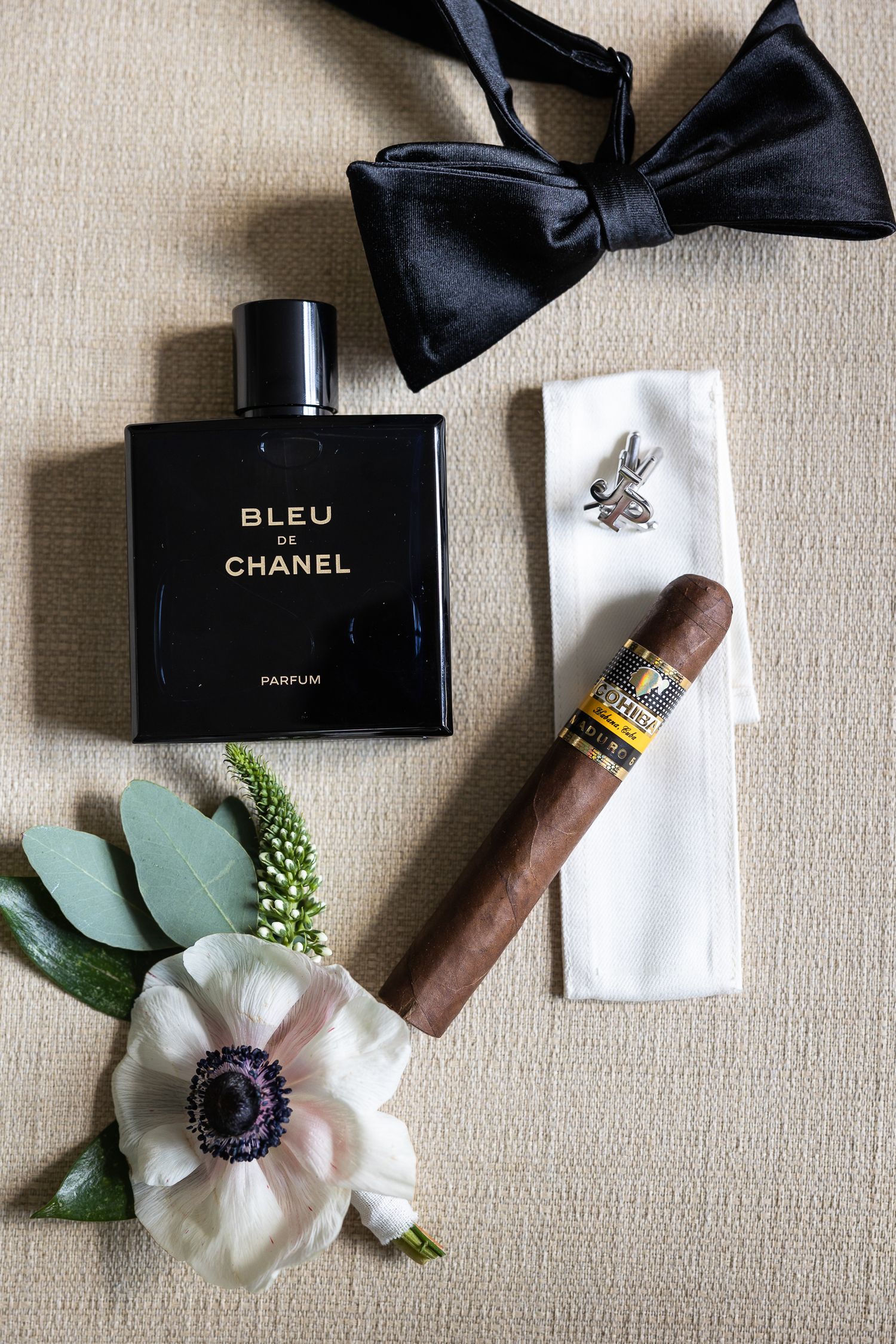 Wedding details including black bow tie, Chanel perfume, cigars and boutonniere on beige background.