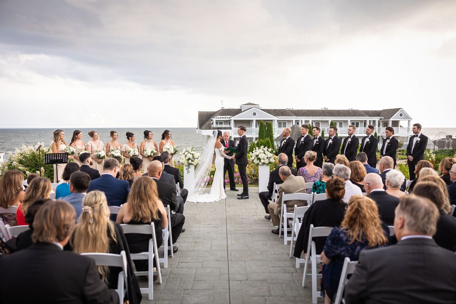 Wedding ceremony guests seated in white chairs on a sunny outdoor patio overlooking water.