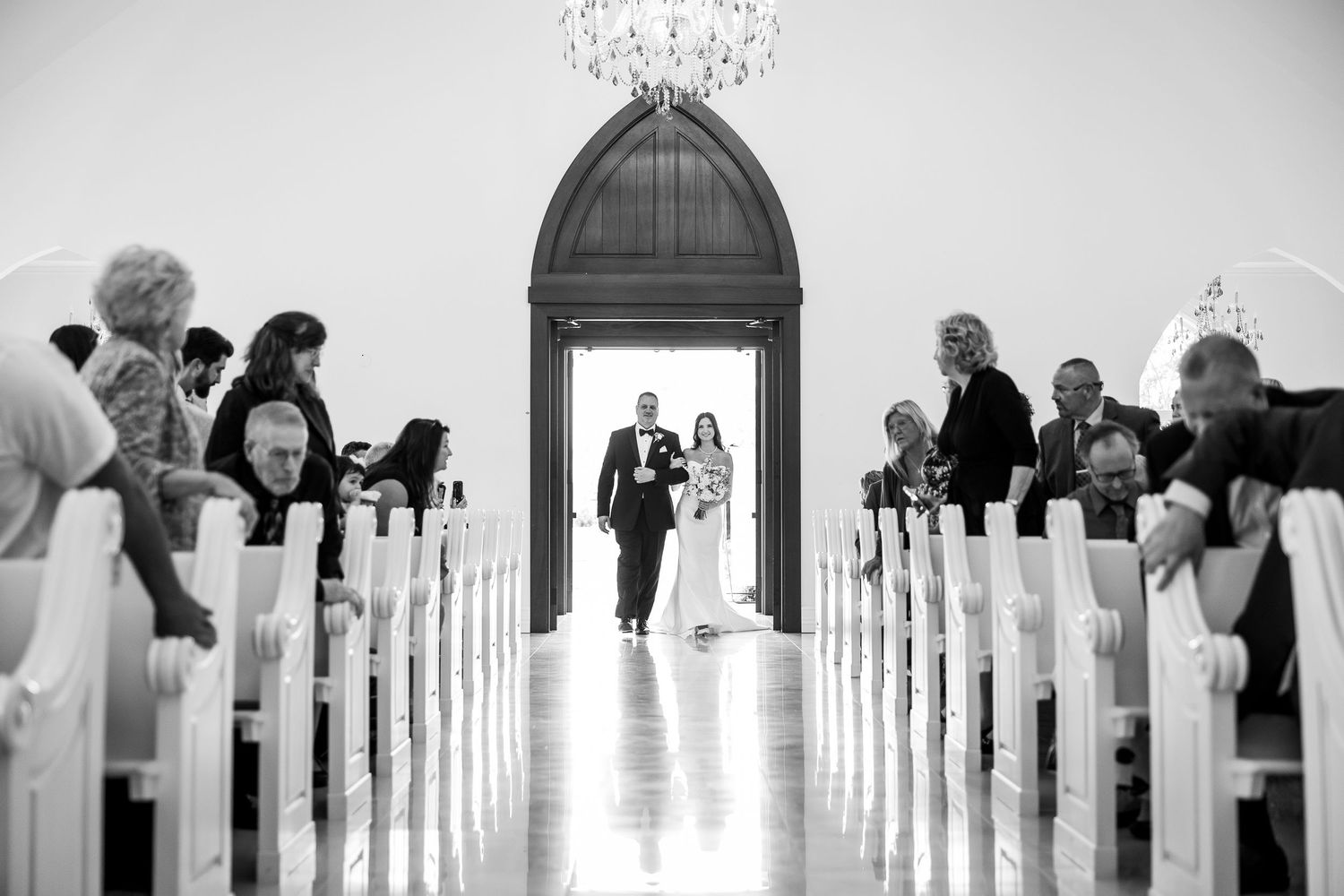 Black and white panorama of church interior with guests seated in pews under chandelier.