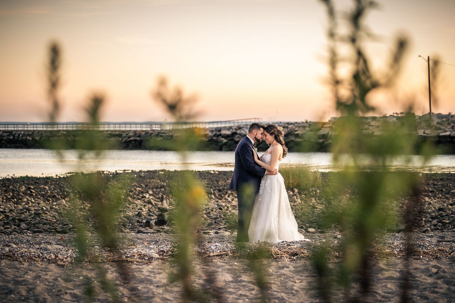 A couple shares a romantic embrace during sunset by a waterfront with mountains in the background.