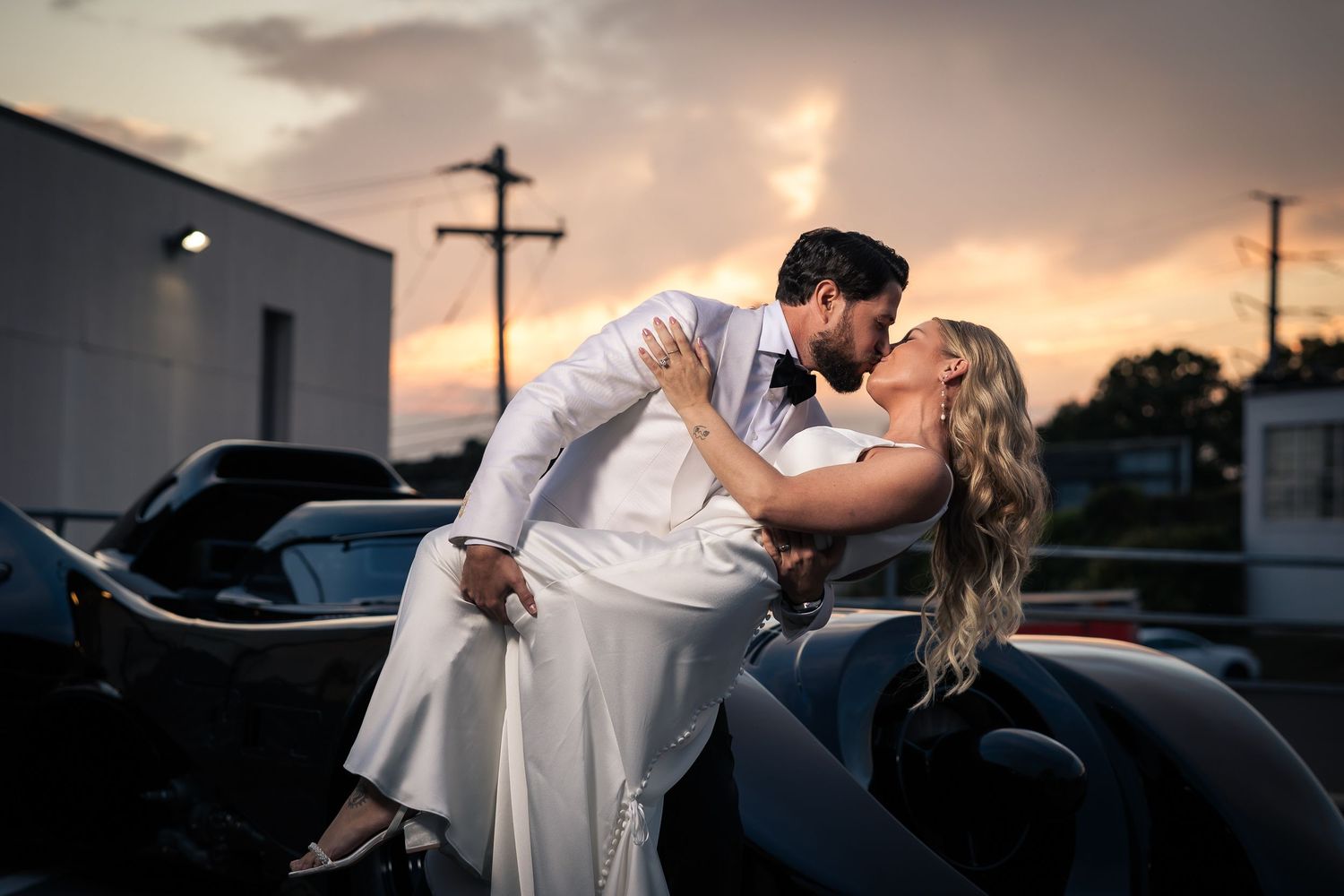 Romantic couple in wedding attire share intimate moments during sunset photoshoot with vintage car.