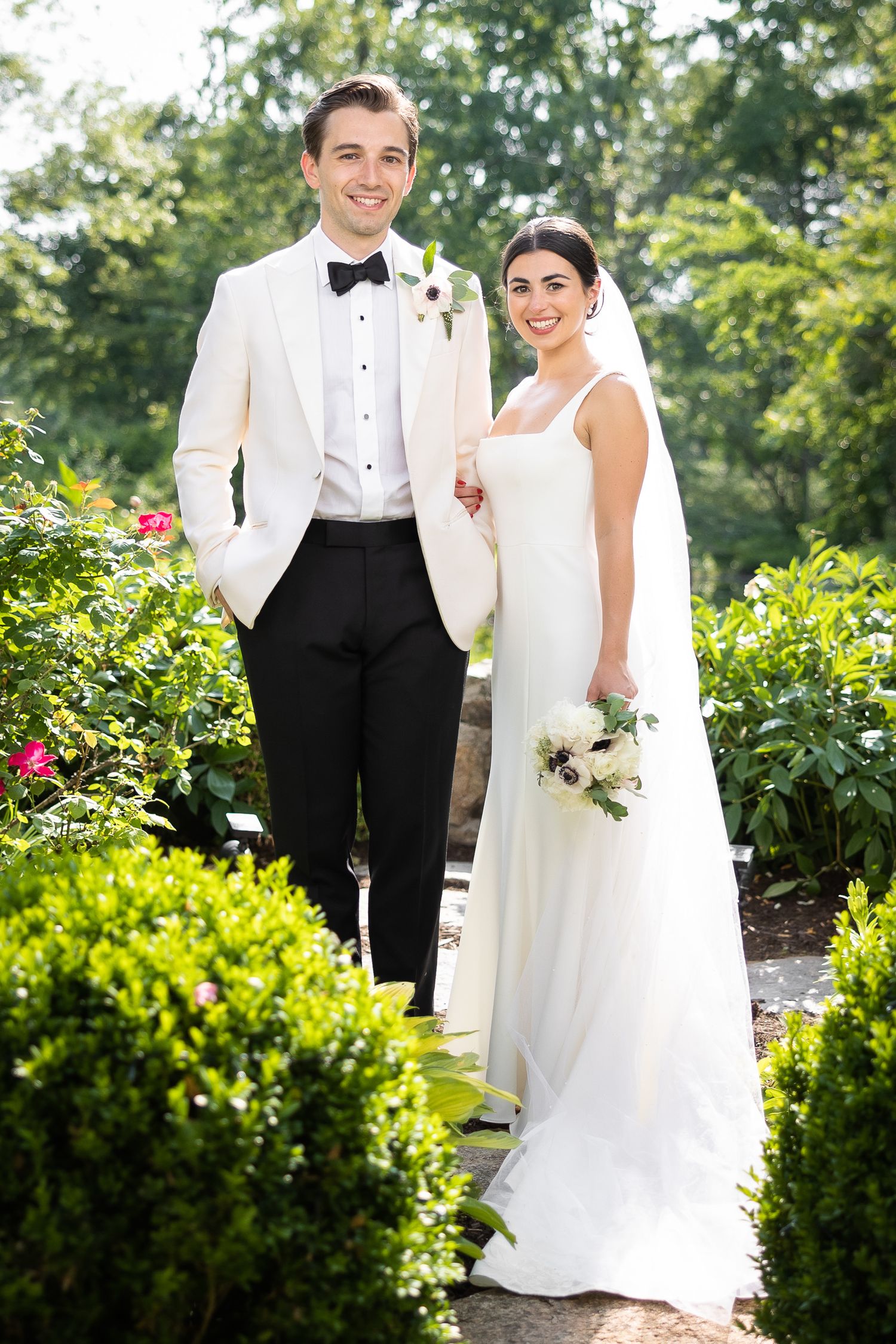 Wedding couple posing in formal attire among blooming garden flowers.