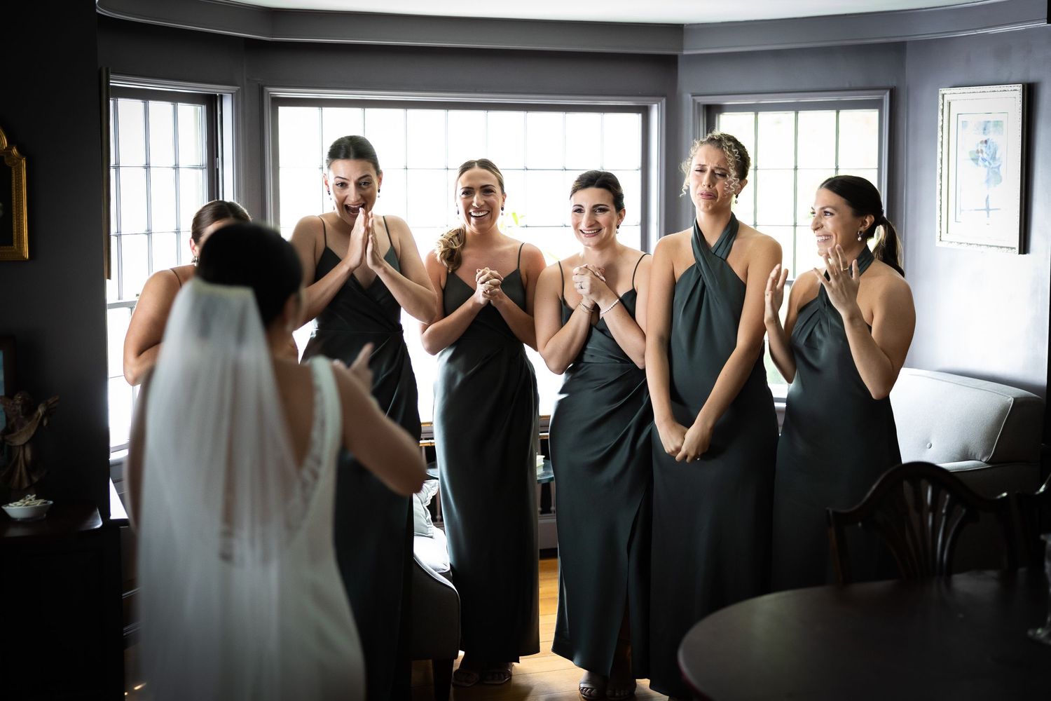 Bridesmaids in dark dresses react emotionally during wedding preparations.