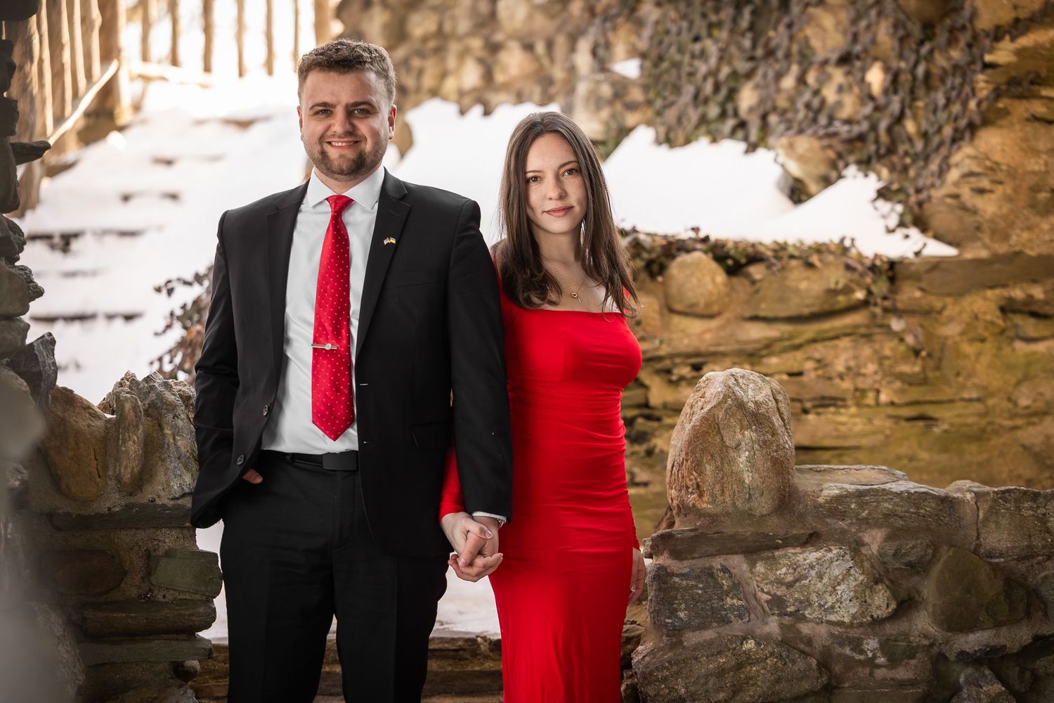 A couple in formal attire poses for romantic photos in front of a snowy rock formation at sunset.