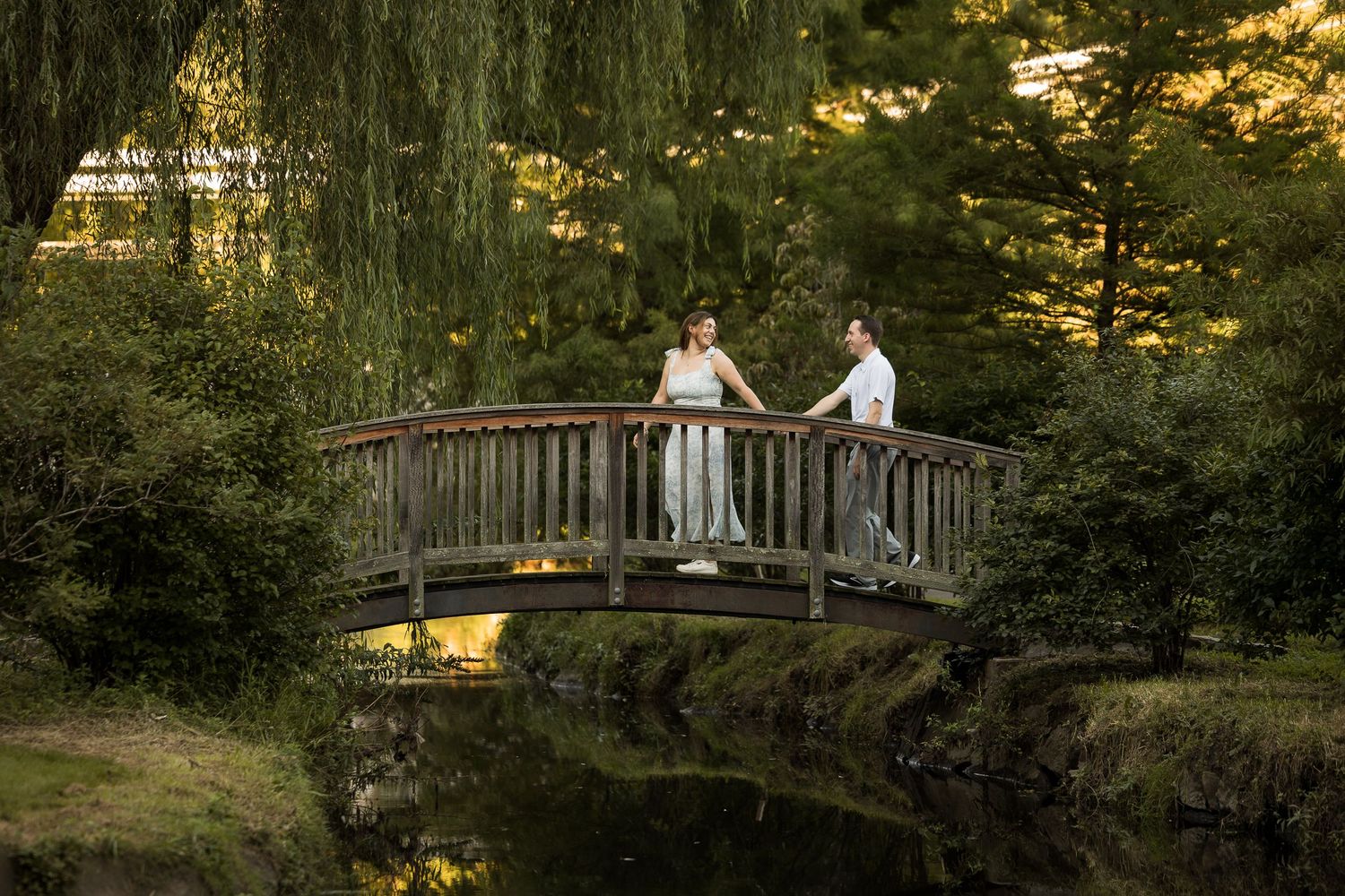 Bride and groom share a romantic moment on a wooden bridge surrounded by lush greenery and a weeping willow at dusk.