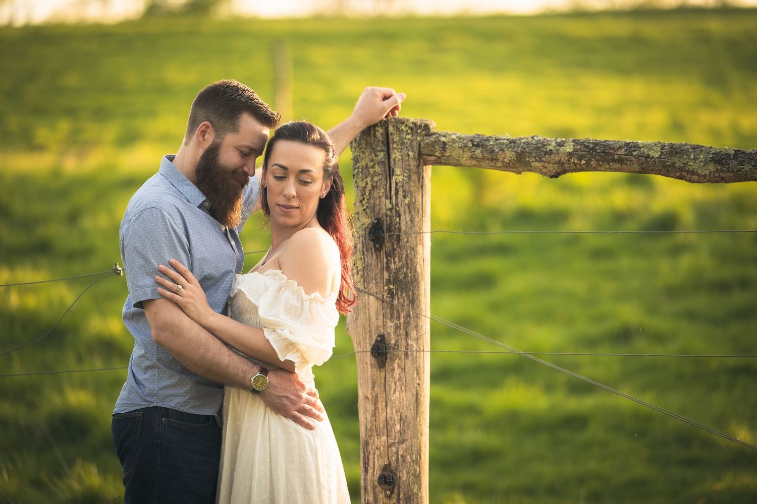 A couple shares an intimate moment together in a sun-drenched meadow near a rustic wooden fence post.