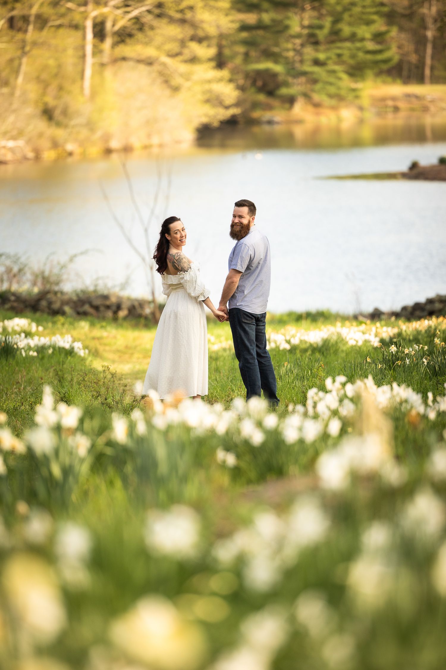 A couple stands together in a meadow of blooming daffodils beside a serene lake on a spring afternoon.