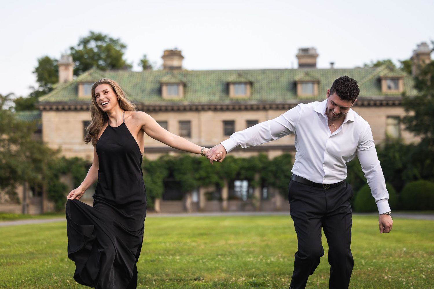A couple in formal attire dances gracefully on a grassy lawn with a historic building in the background.