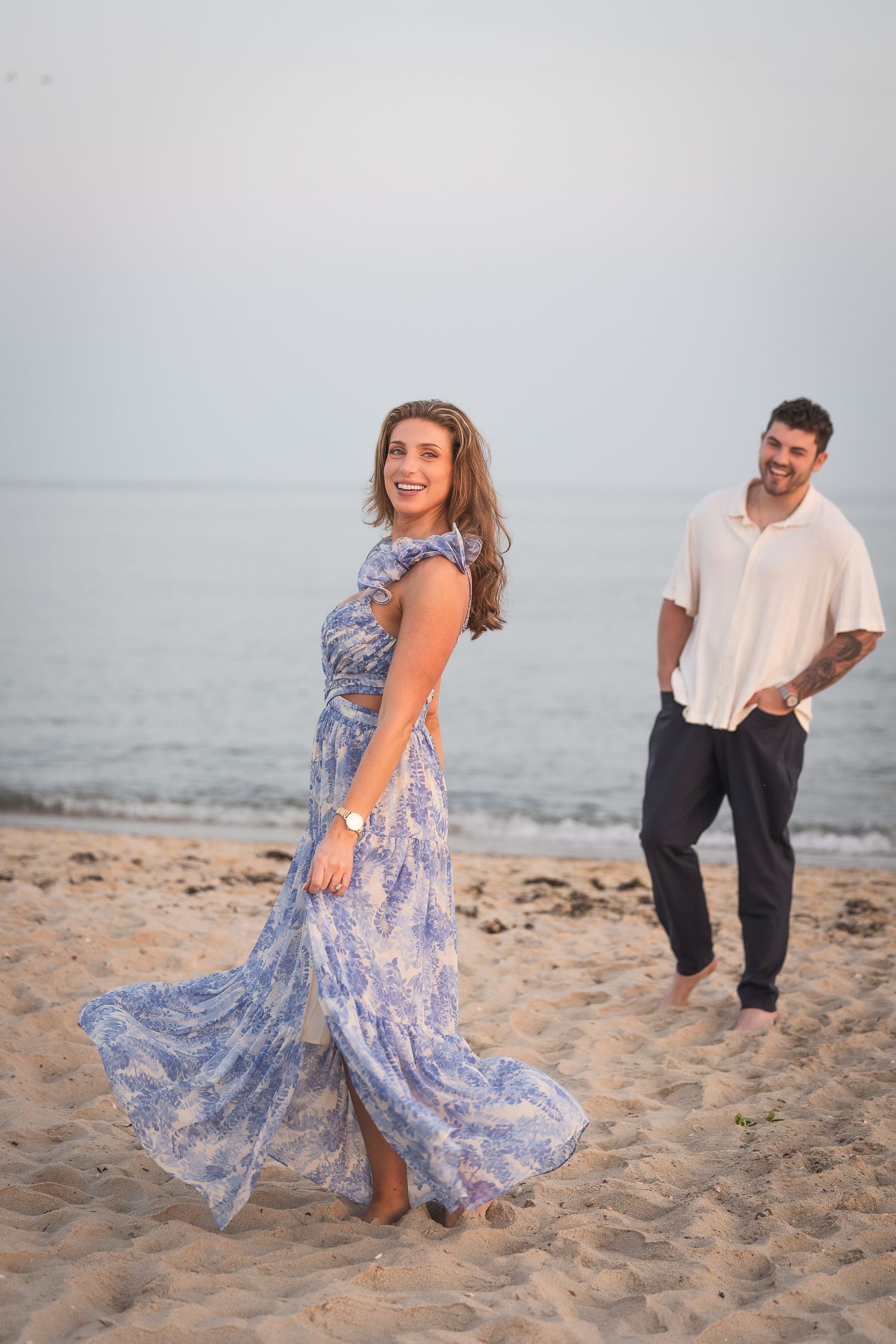 A couple shares a playful moment together on a misty beach while wearing flowy blue dress and casual attire.