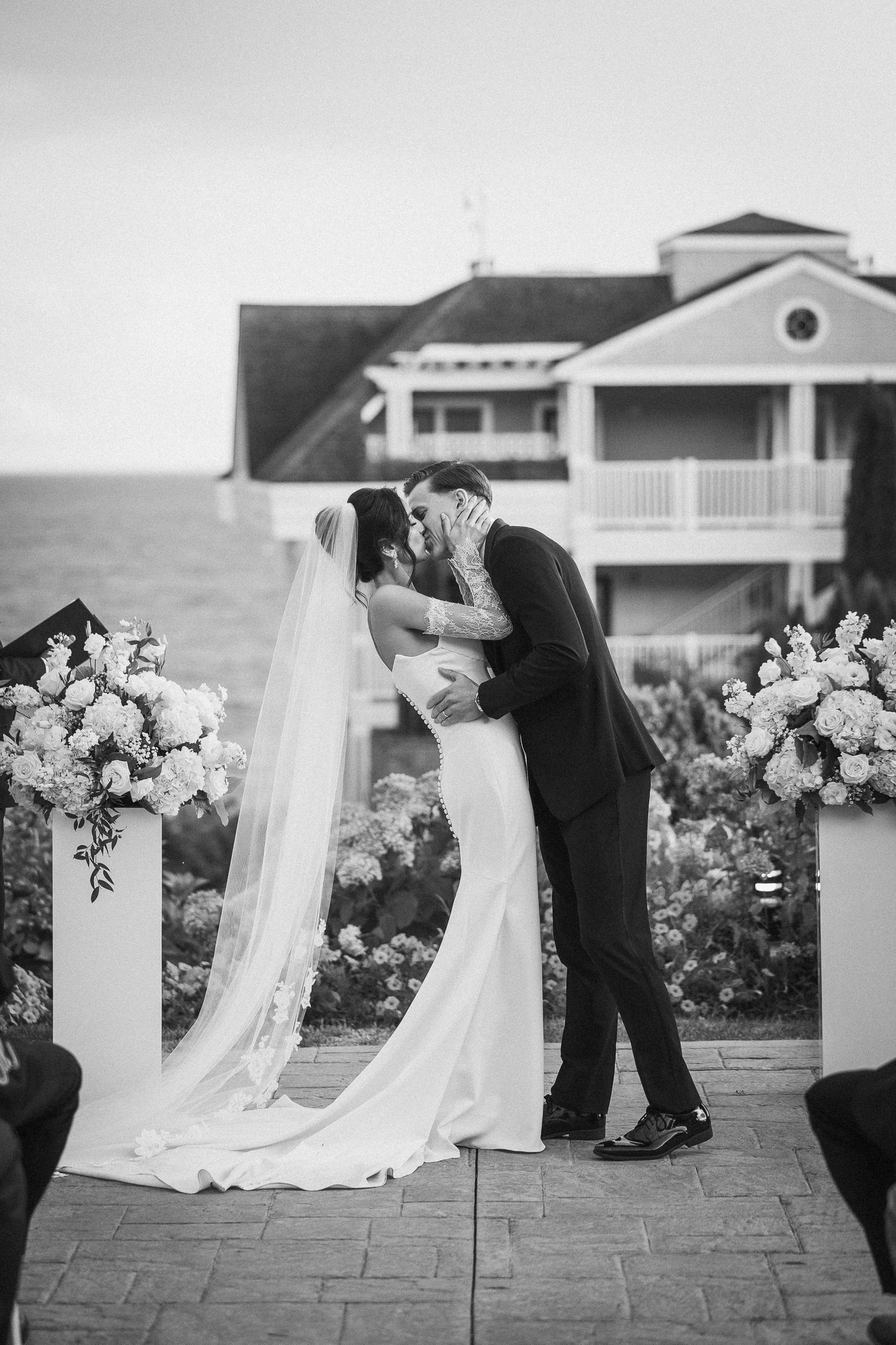 Romantic wedding ceremony kiss with floral arrangements and coastal house backdrop.