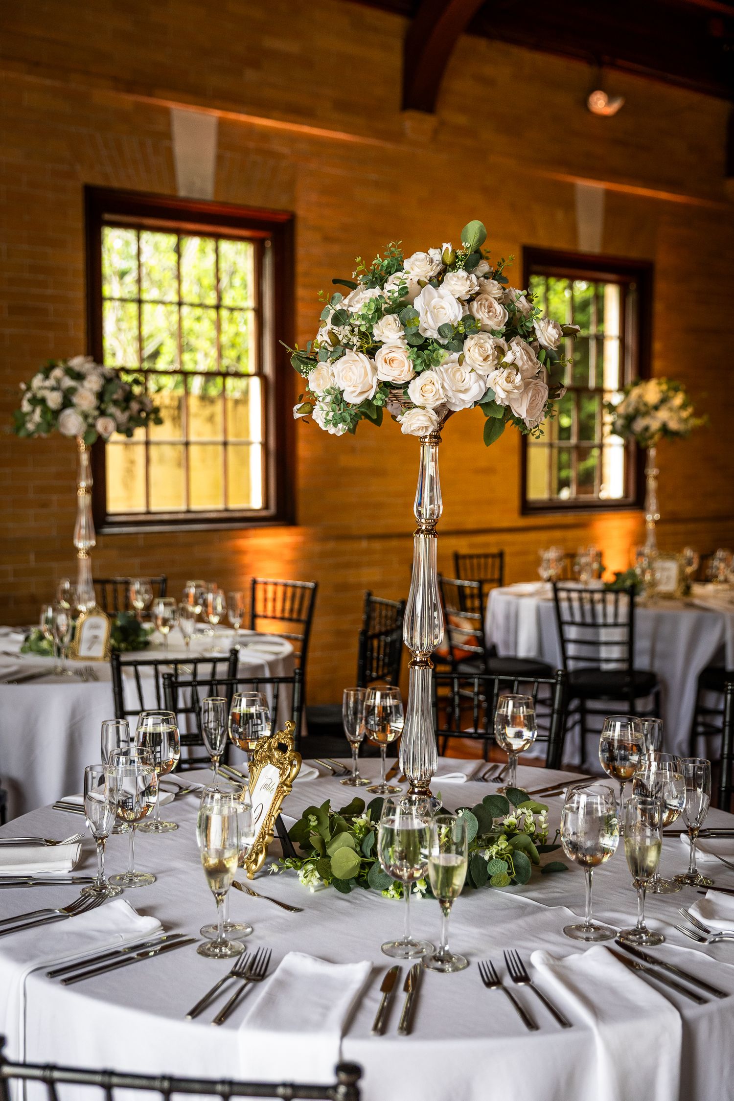Elegant wedding reception tables with white linens and tall floral centerpieces in rustic venue.