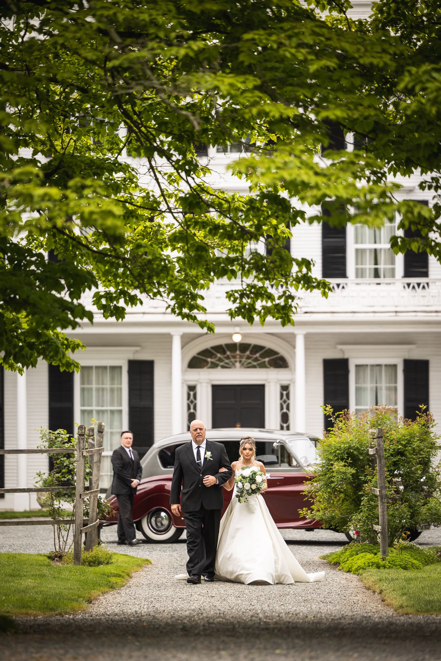 Wedding party poses in front of classic white colonial mansion with vintage car.