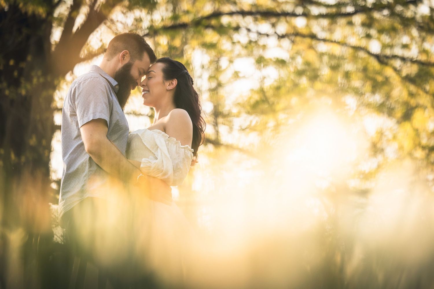 Silhouetted couple embraces in a golden sunset with sunbeams streaming through trees in a romantic outdoor setting.