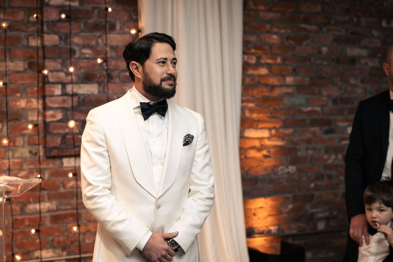 A group in white tuxedos poses against a rustic brick wall with string lights in an elegant setting.