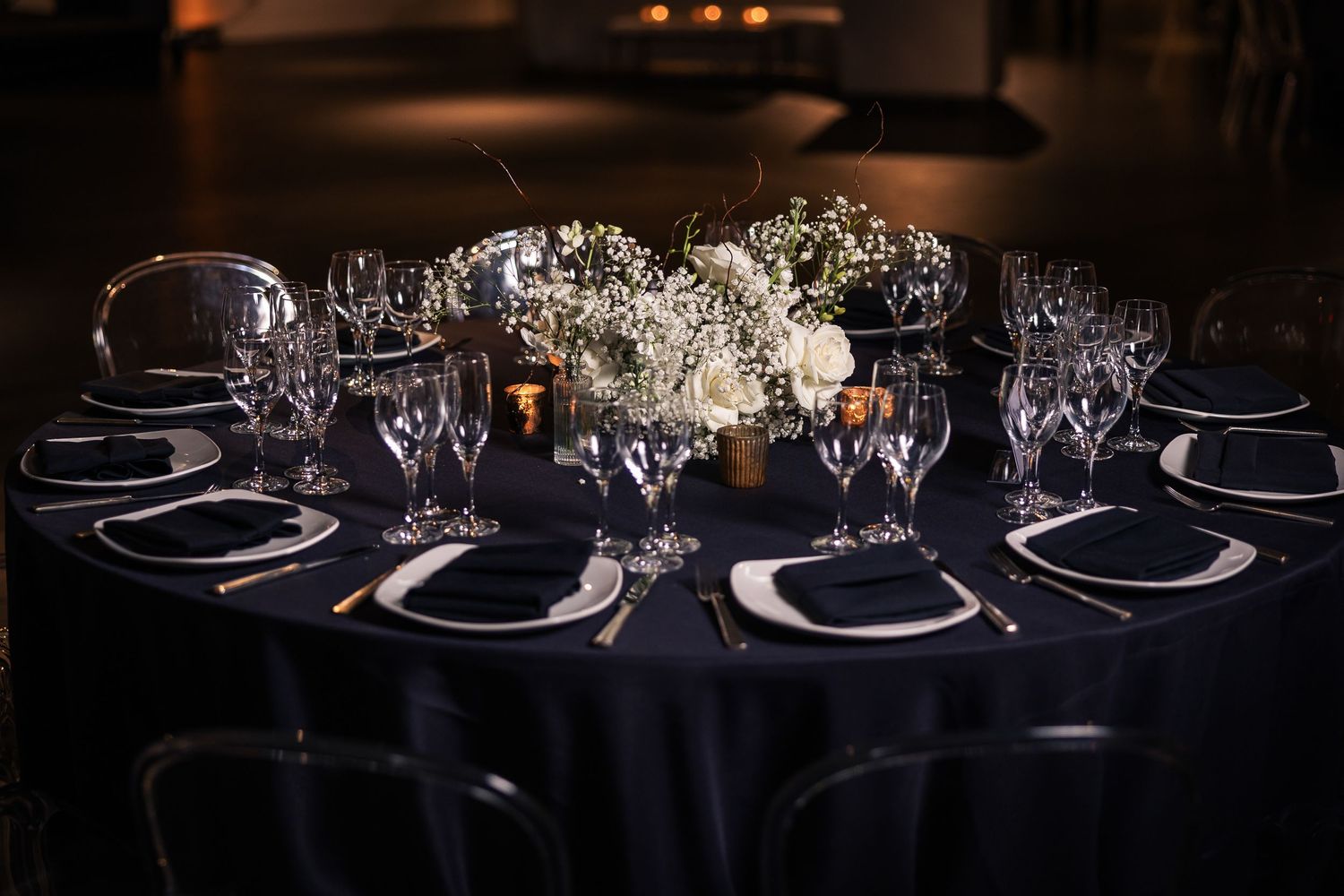 Elegant table settings with dark navy blue tablecloths and white place settings in a dimly lit event space.