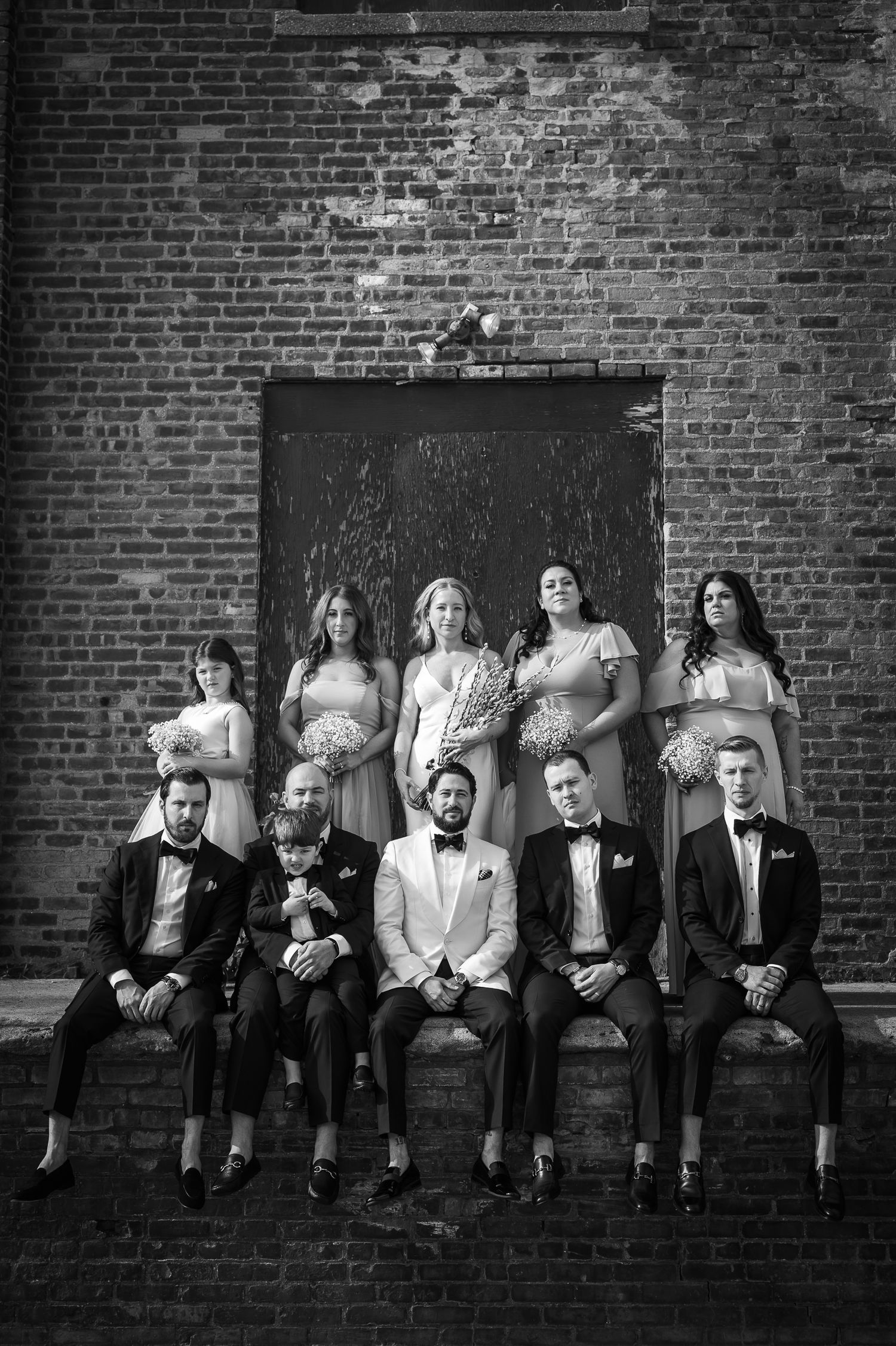 Black and white wedding party portrait with bridesmaids standing and groomsmen seated against rustic brick wall.