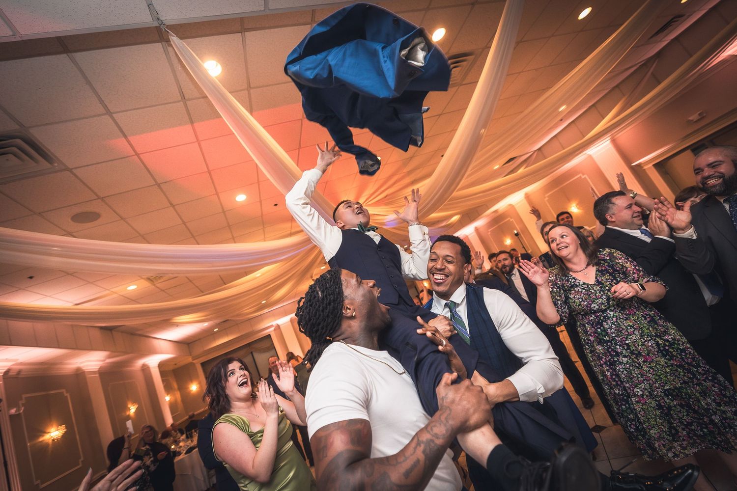 Wedding guests dance and celebrate on a dimly lit dance floor with draped fabric ceiling decorations and party lighting.