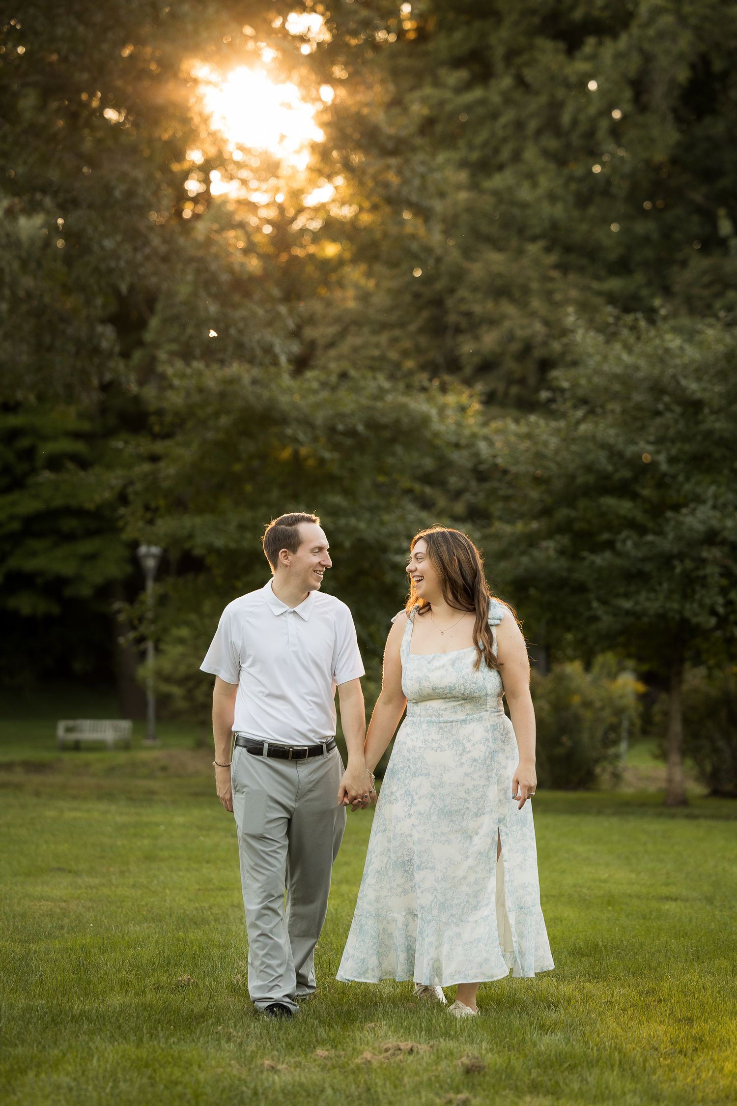Couple holding hands and smiling while walking on green grass at golden hour with sunlit trees in background.