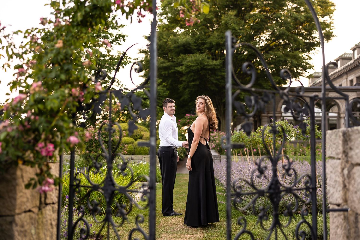 A couple poses in formal attire against an ornate iron gate with flowering trees in the background.