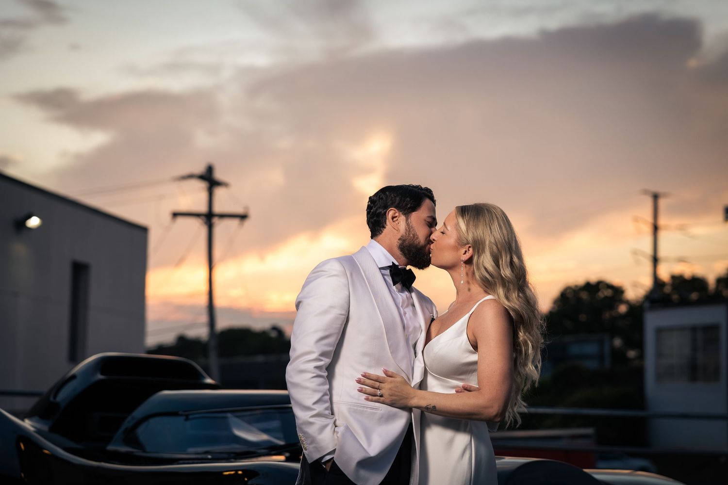 A romantic couple shares a tender kiss at sunset with a dramatic sky and silhouetted buildings in the background.