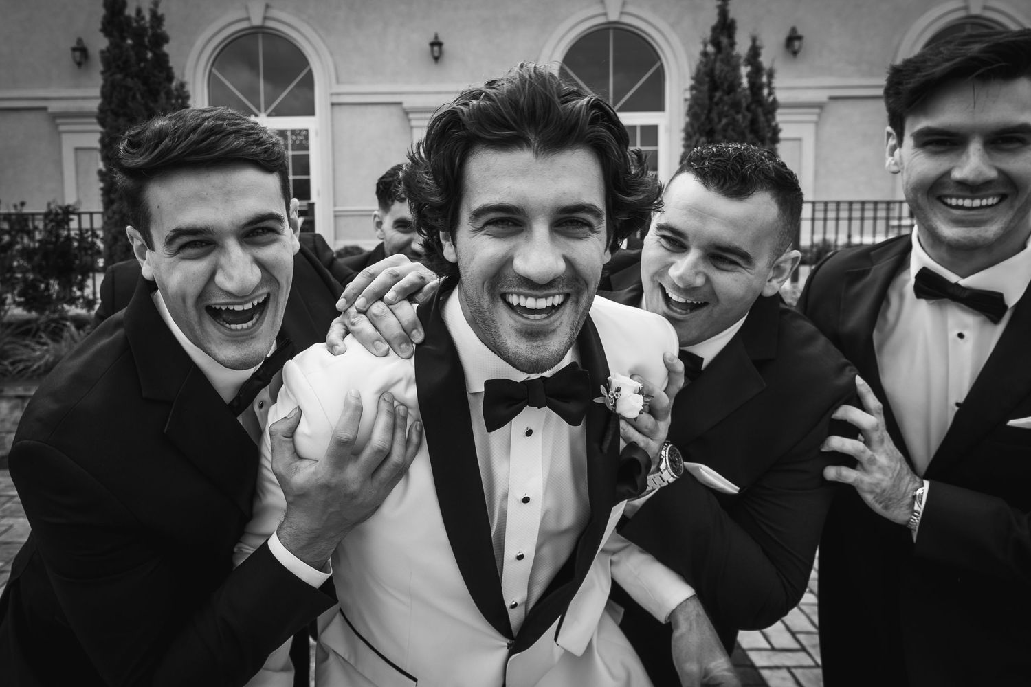 Group of groomsmen in tuxedos laughing and celebrating together in black and white.