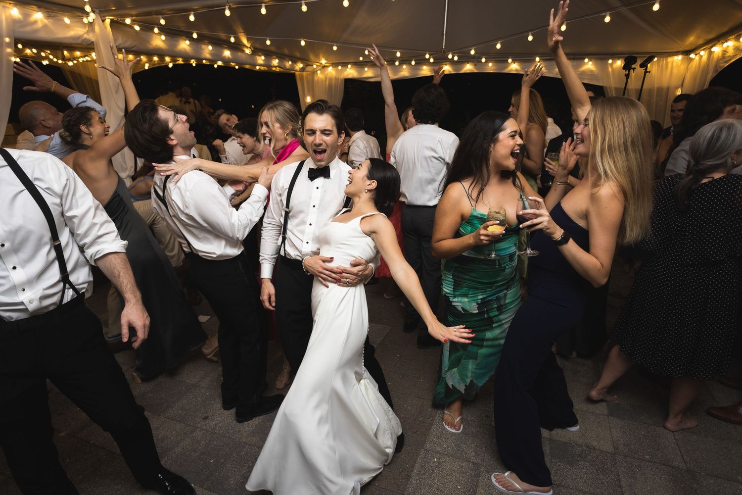 Wedding reception dance floor filled with guests under string lights at night.