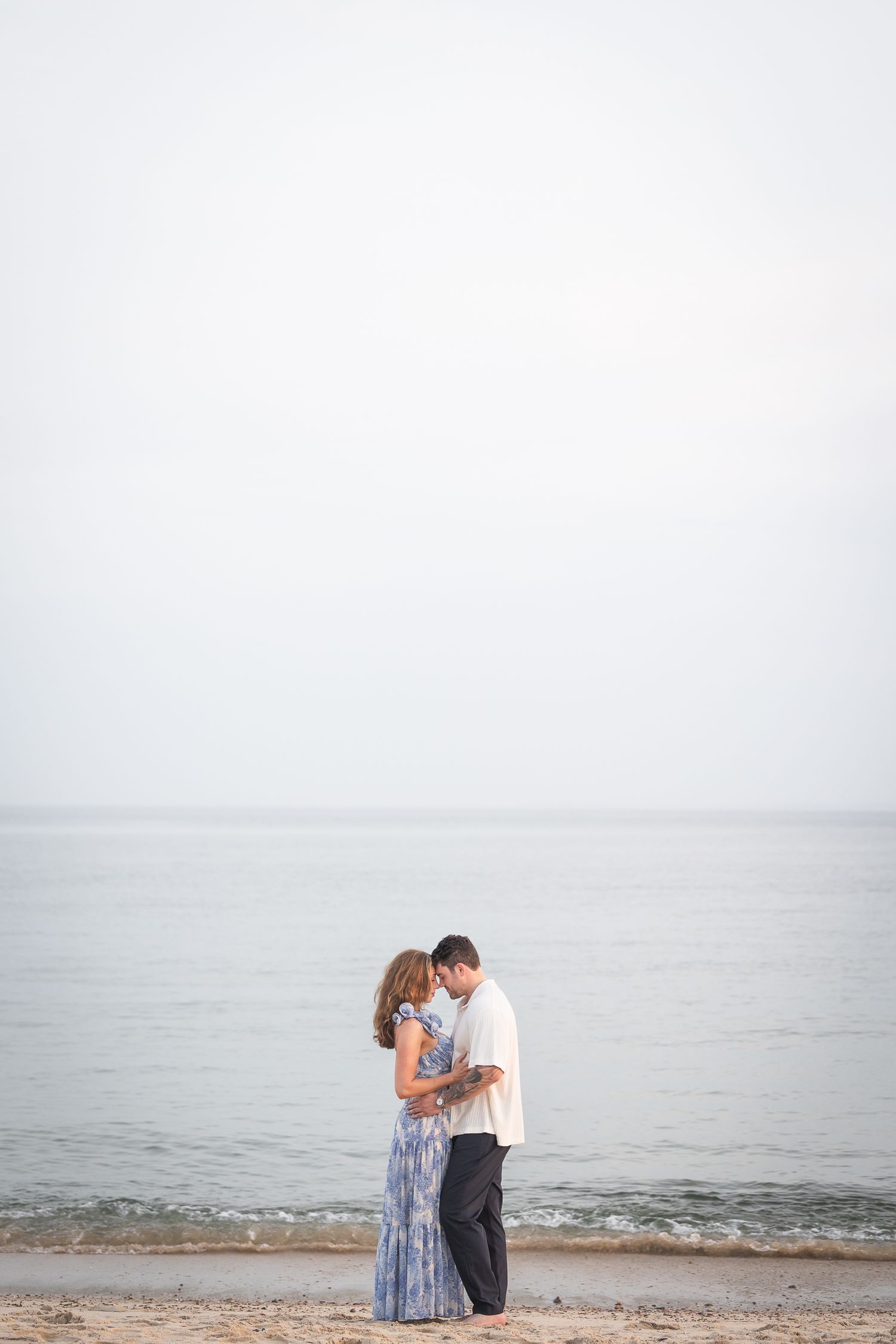 A couple embraces on a misty beach shore with calm ocean waters and soft gray skies in the background.