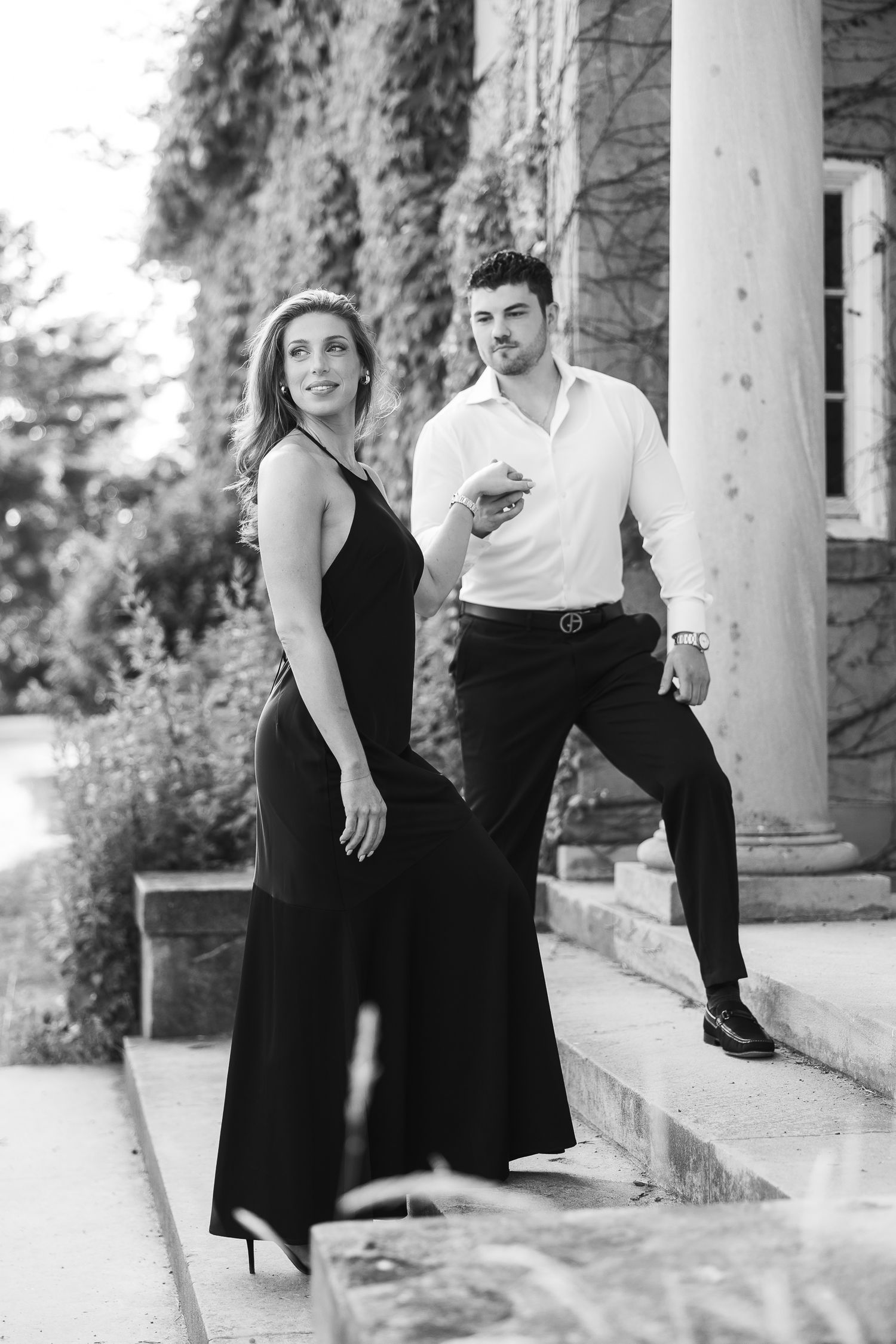 A couple in formal black and white attire poses together on stone steps in an elegant outdoor setting.