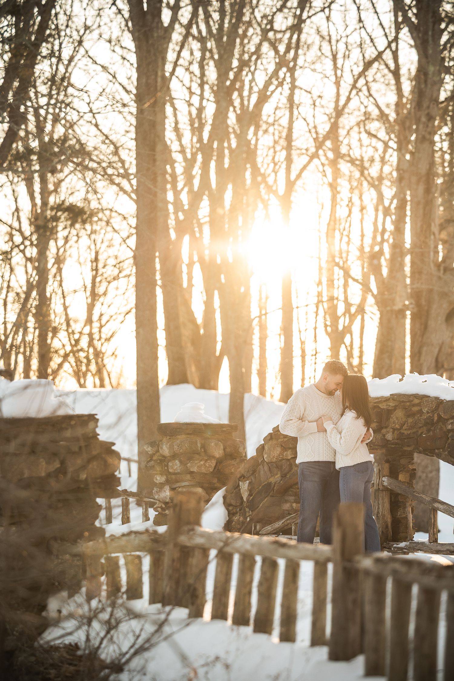 Couple walking through snow-covered woods during golden hour with sunlight streaming through bare winter trees.