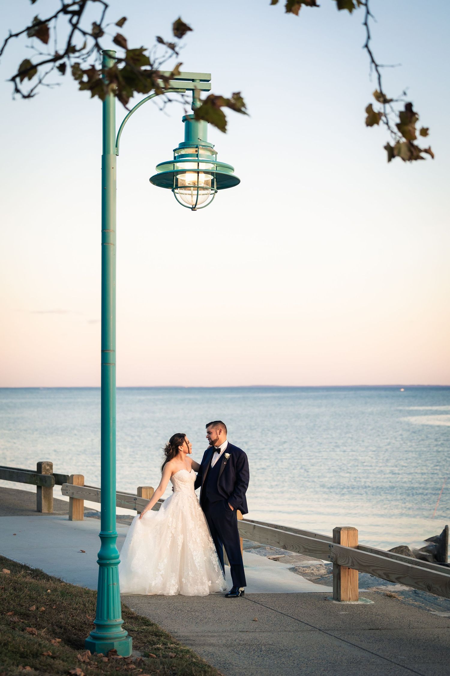 A couple in wedding attire shares romantic moments along a coastal boardwalk with turquoise lamp posts at sunset.