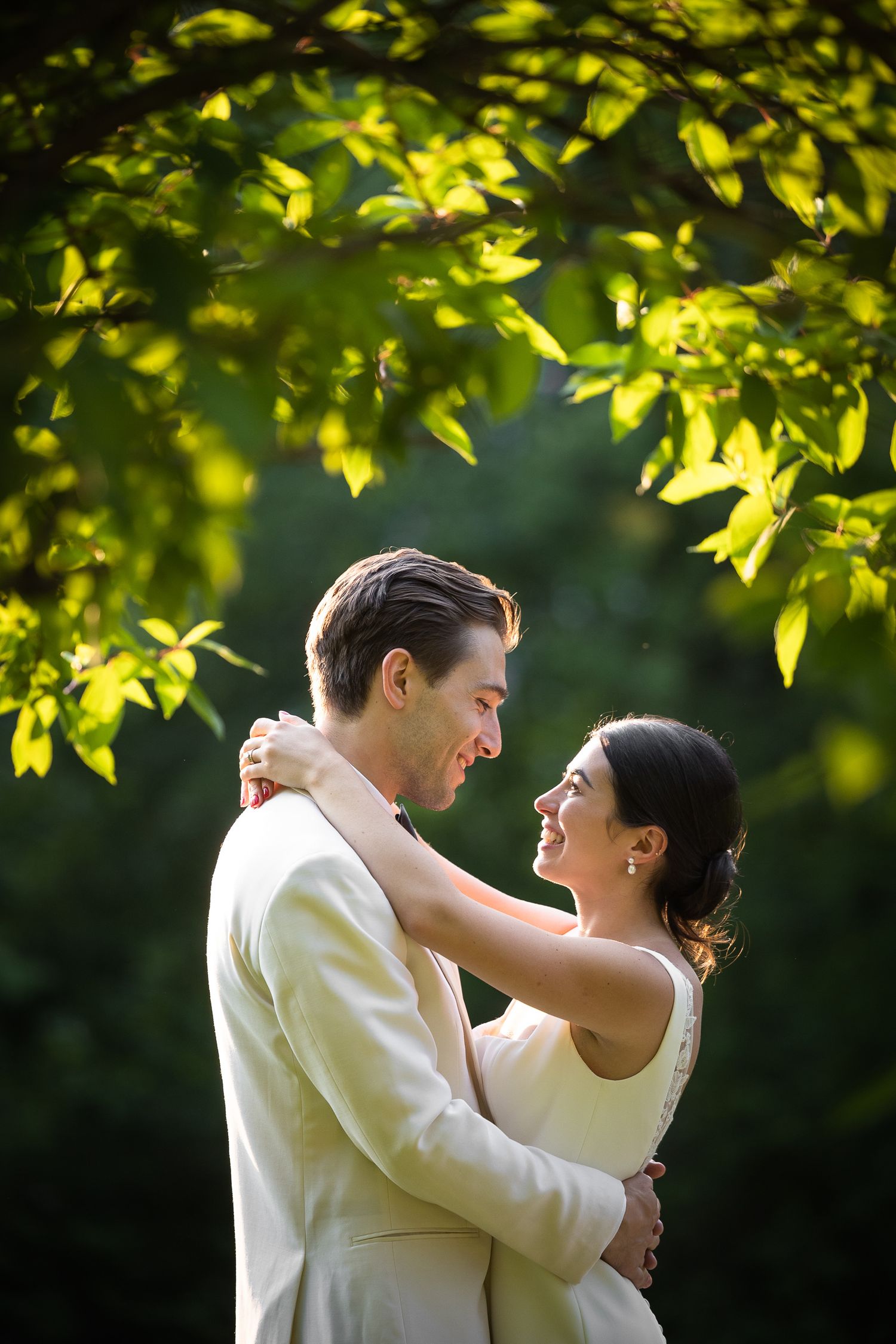 Joyful couple embraces under sunlit green leaves during outdoor wedding celebration.
