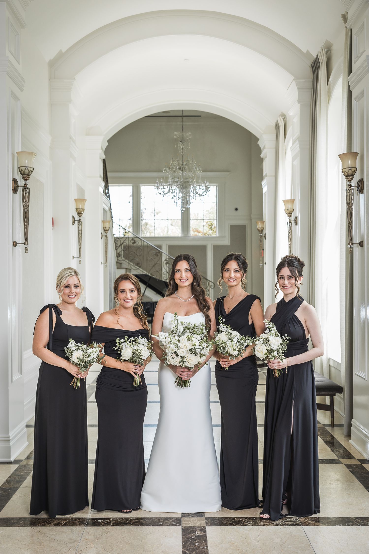 Wedding party poses in elegant white hallway with bridesmaids in black dresses holding bouquets.