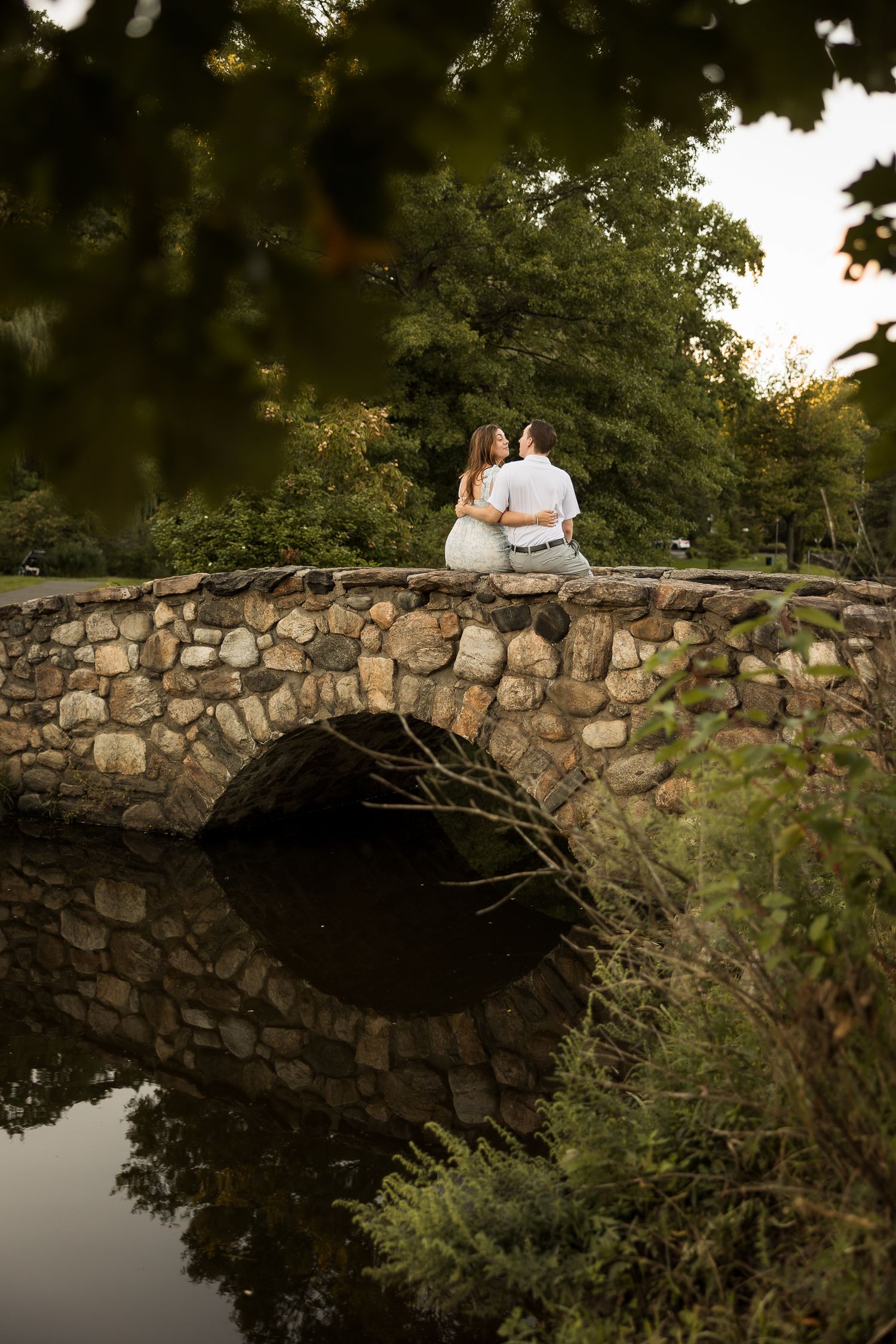 Couple sitting closely on a rustic stone arch bridge reflected in calm water, surrounded by lush green trees.