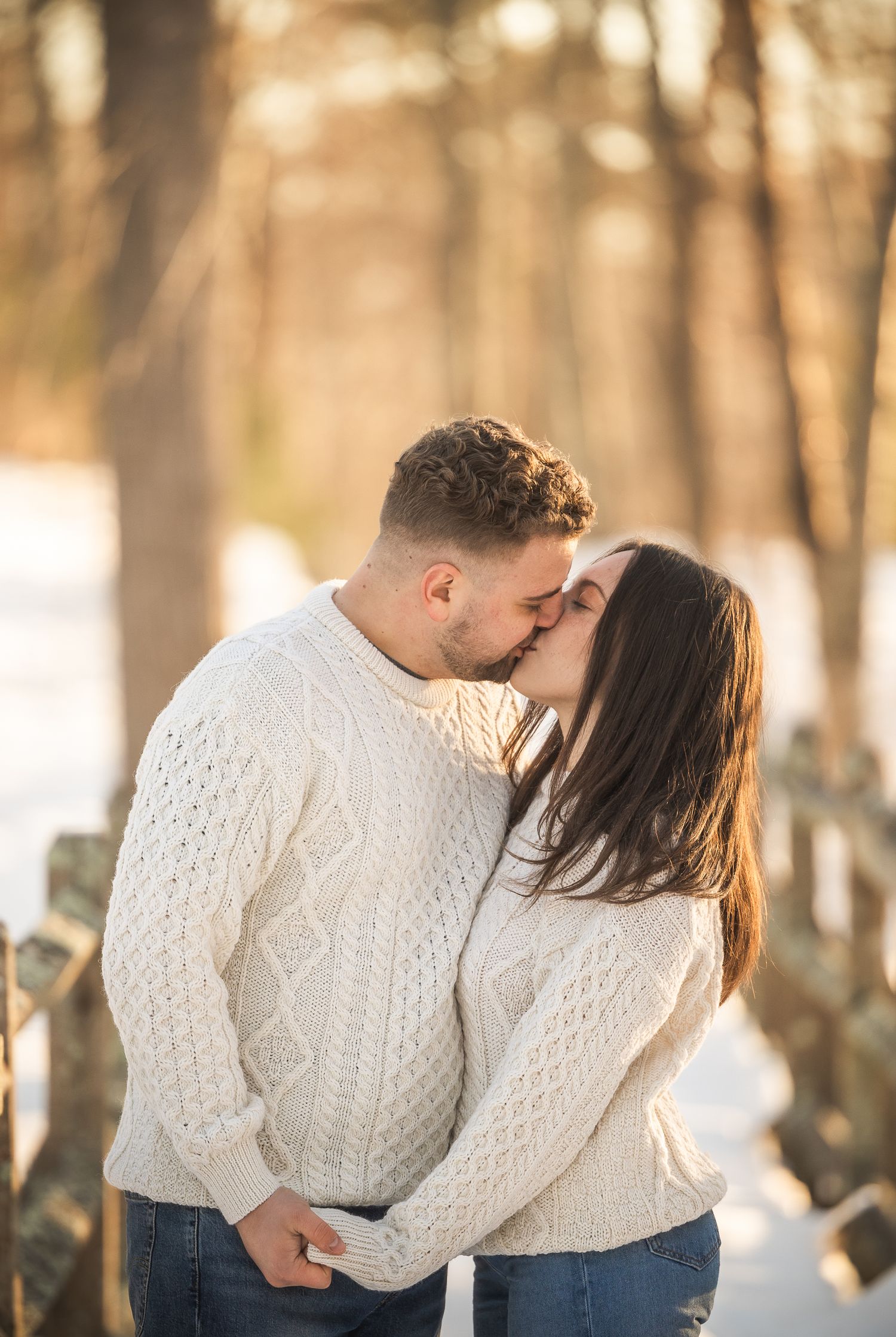 A romantic couple in matching white sweaters embrace in a sunlit forest setting during an autumn photoshoot.