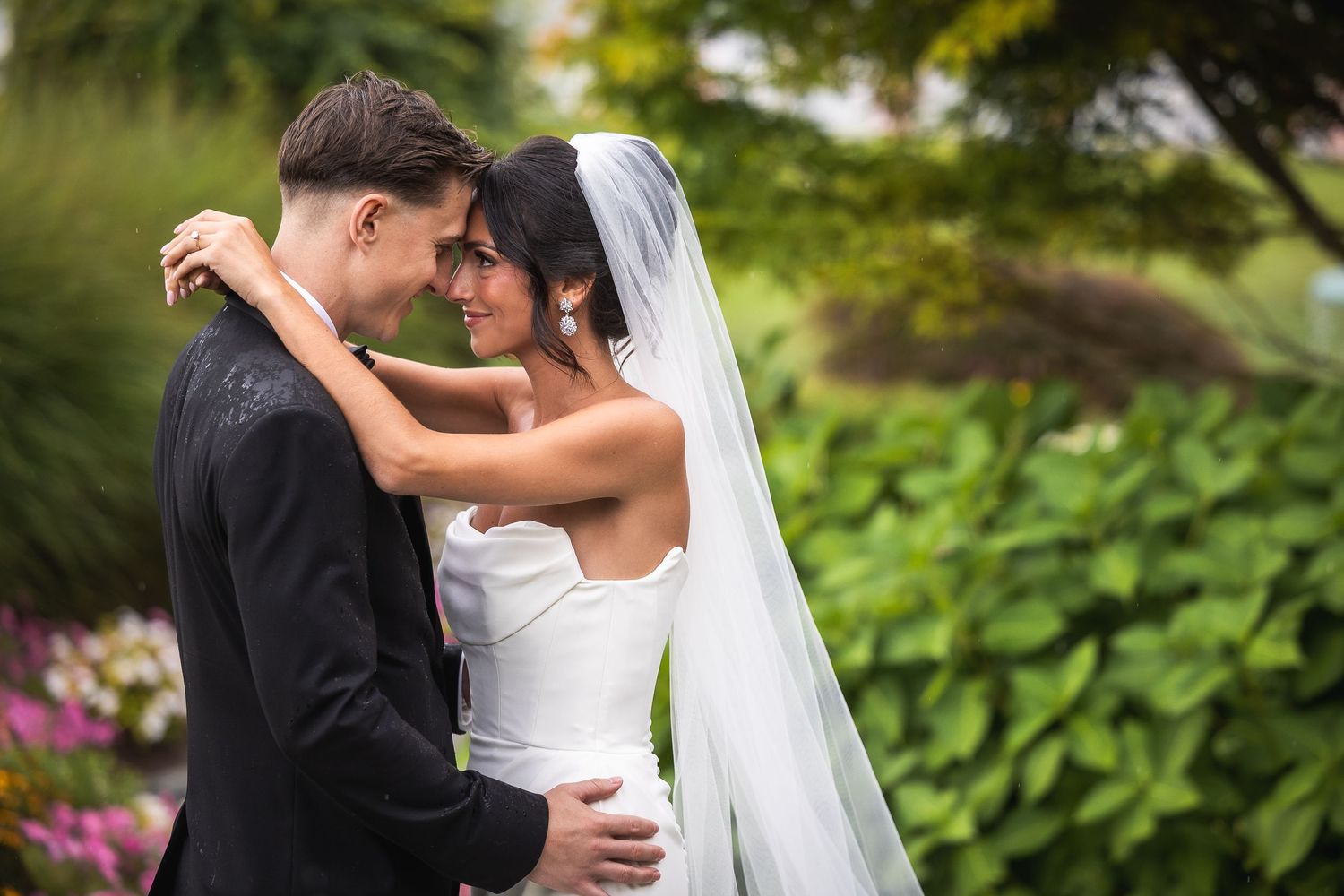 Couple shares romantic kiss in garden setting with greenery backdrop.